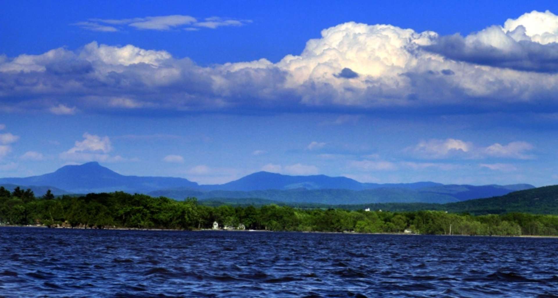 A serene lake surrounded by lush green forests, with majestic mountains rising in the distance against a vibrant blue sky dotted with fluffy white clouds.