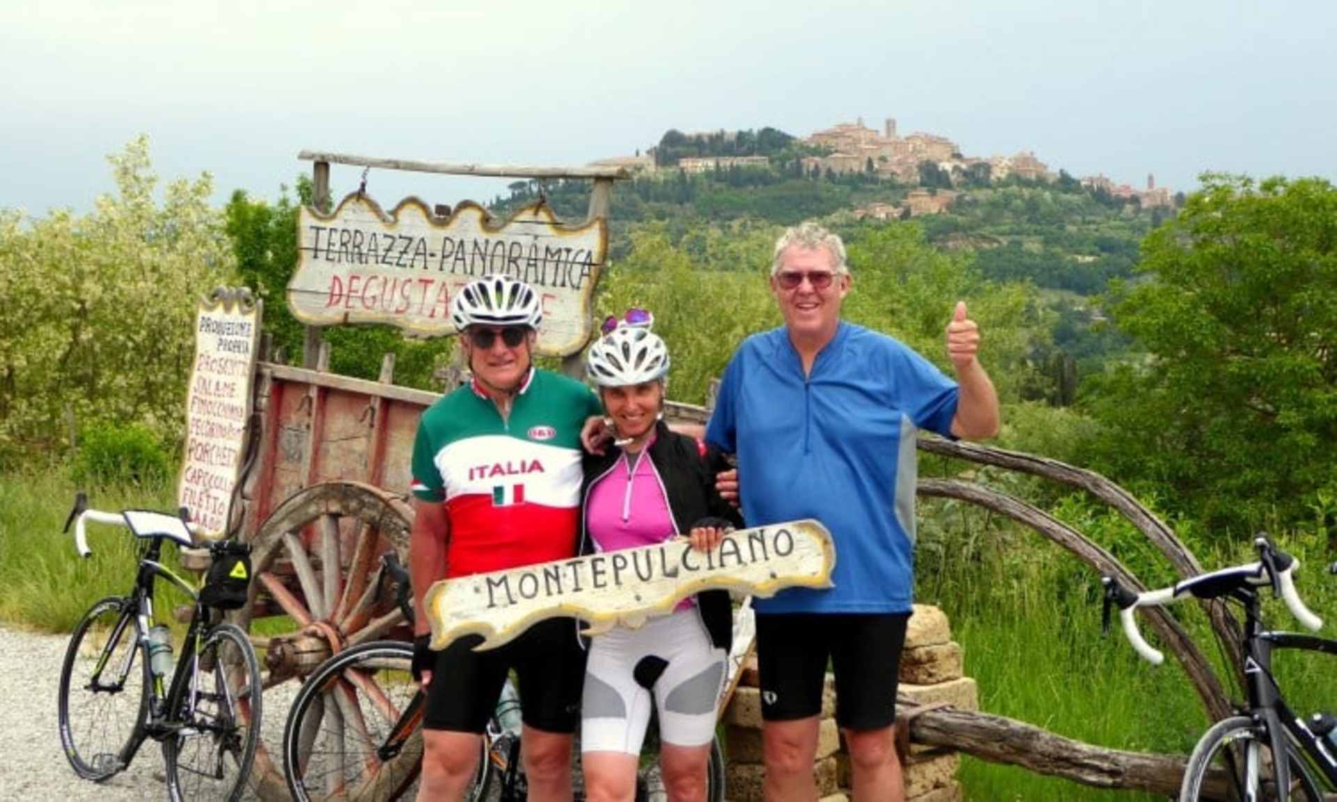 Two cyclists, a man and a woman, posing in front of a scenic rural landscape with a medieval hilltop town in the background.