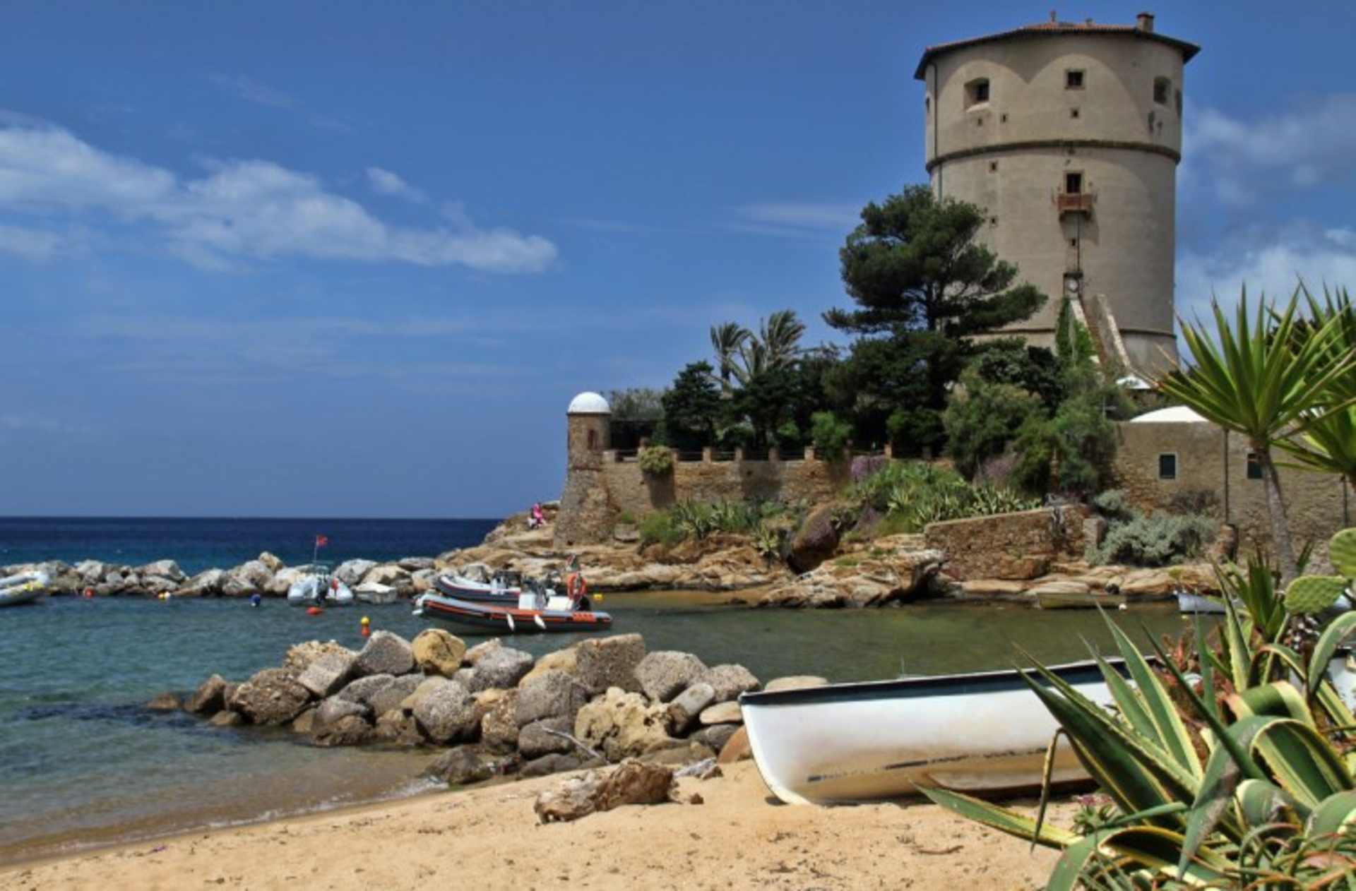A picturesque coastal tower surrounded by rocky shores, palm trees, and a small boat in the foreground, set against a vibrant blue sky with fluffy clouds.