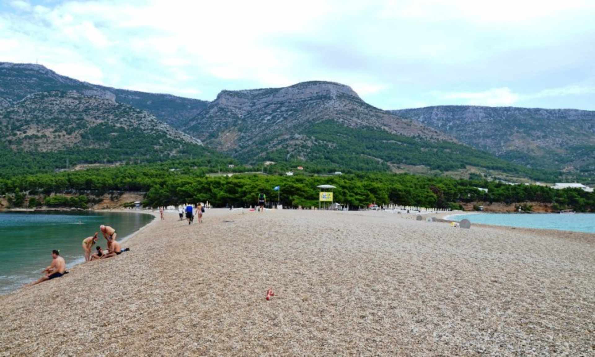 A sandy beach stretches out in the foreground, with people enjoying the sunny day, while the background features a mountainous landscape with lush greenery.