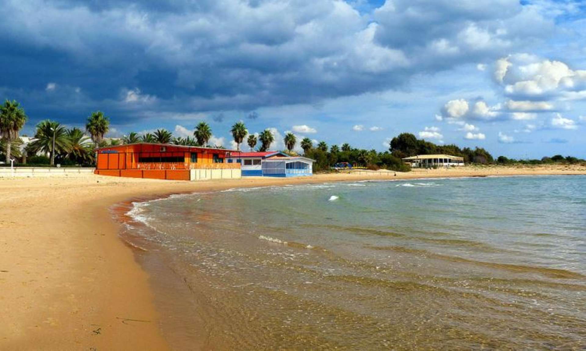 A scenic beach with colorful buildings and palm trees along the shore, set against a dramatic cloudy sky.