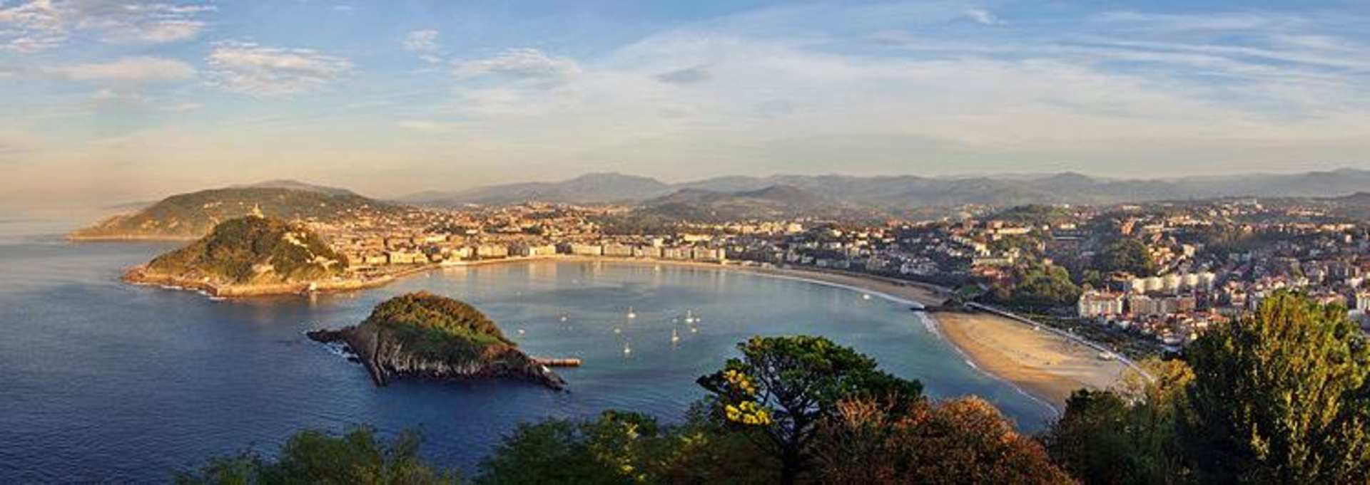 A picturesque coastal town nestled between mountains, with a crescent-shaped bay and boats dotting the turquoise waters in the foreground.
