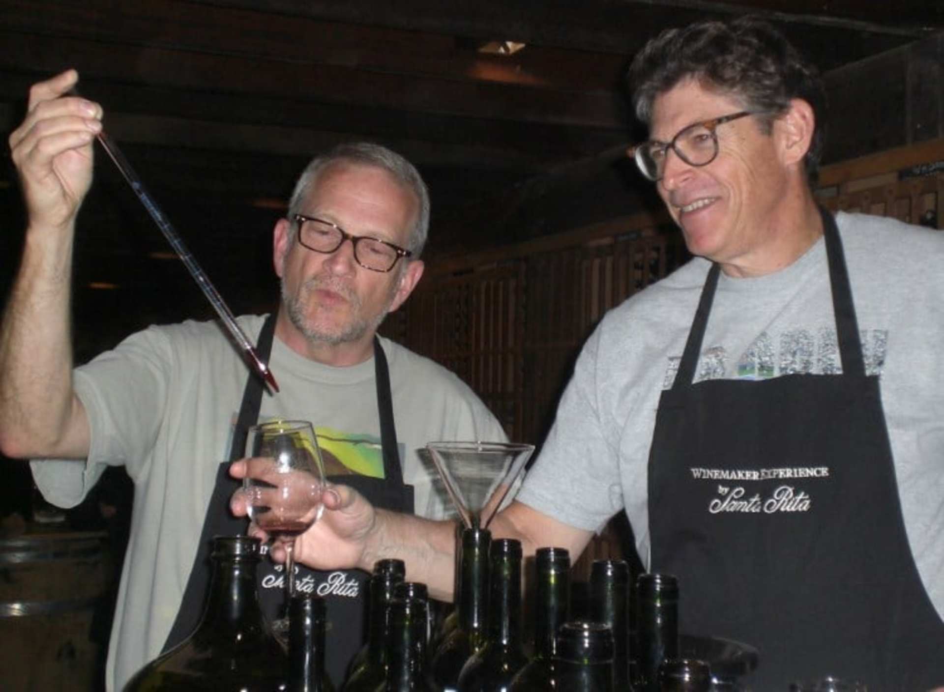 Two men wearing aprons and glasses are standing behind a bar, one holding a glass and the other a bottle, appearing to be engaged in conversation or preparing drinks.