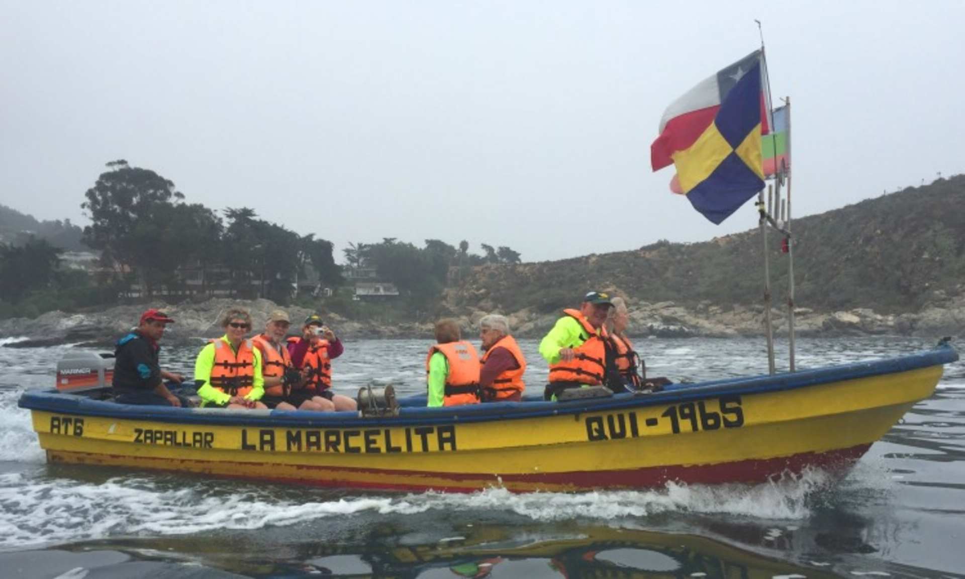 A yellow boat with several people wearing life jackets is navigating through the water, with a mountainous landscape and a colorful flag visible in the background.