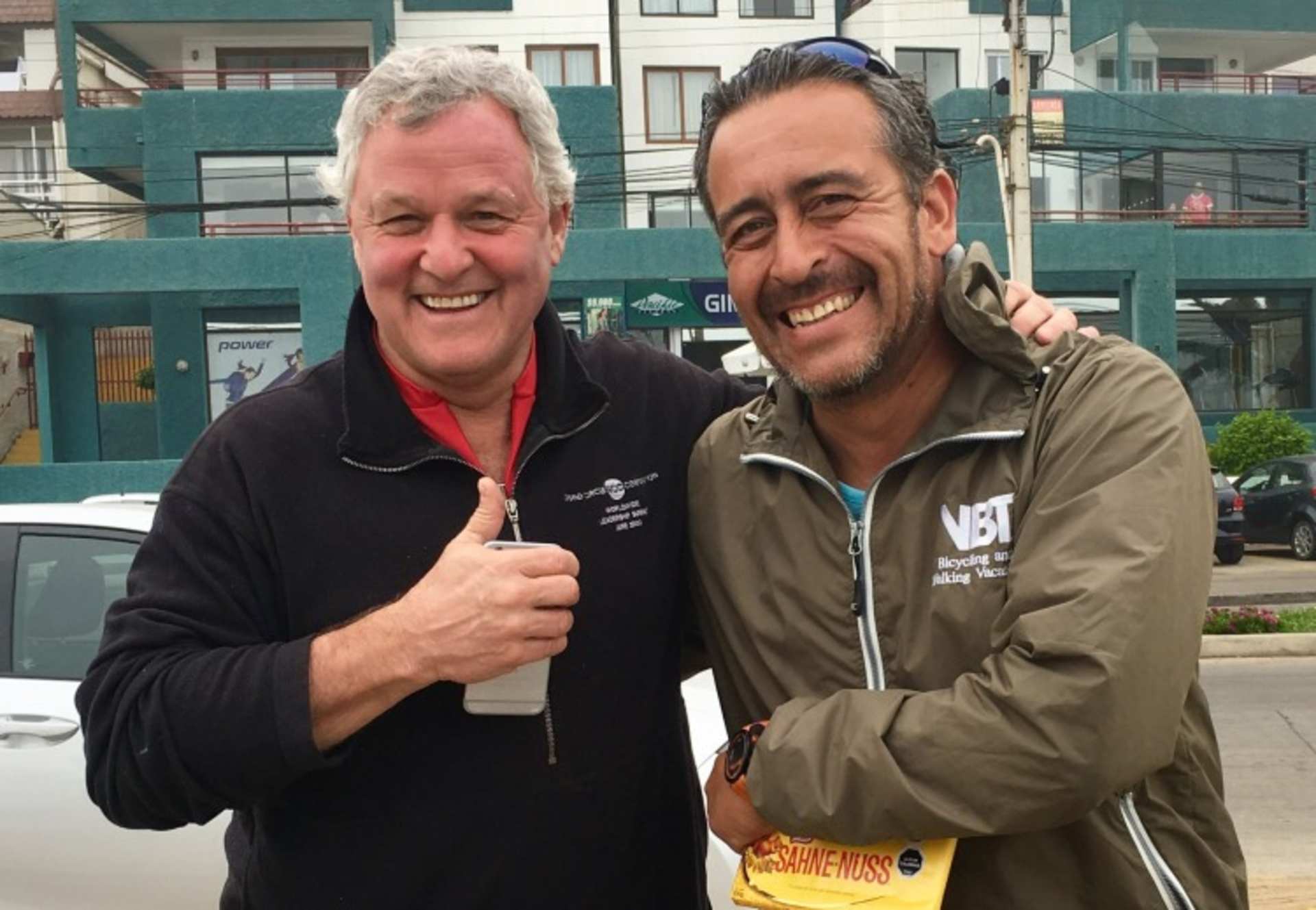 Two men, one with gray hair and the other with dark hair, are standing together and smiling in an urban setting with buildings in the background.