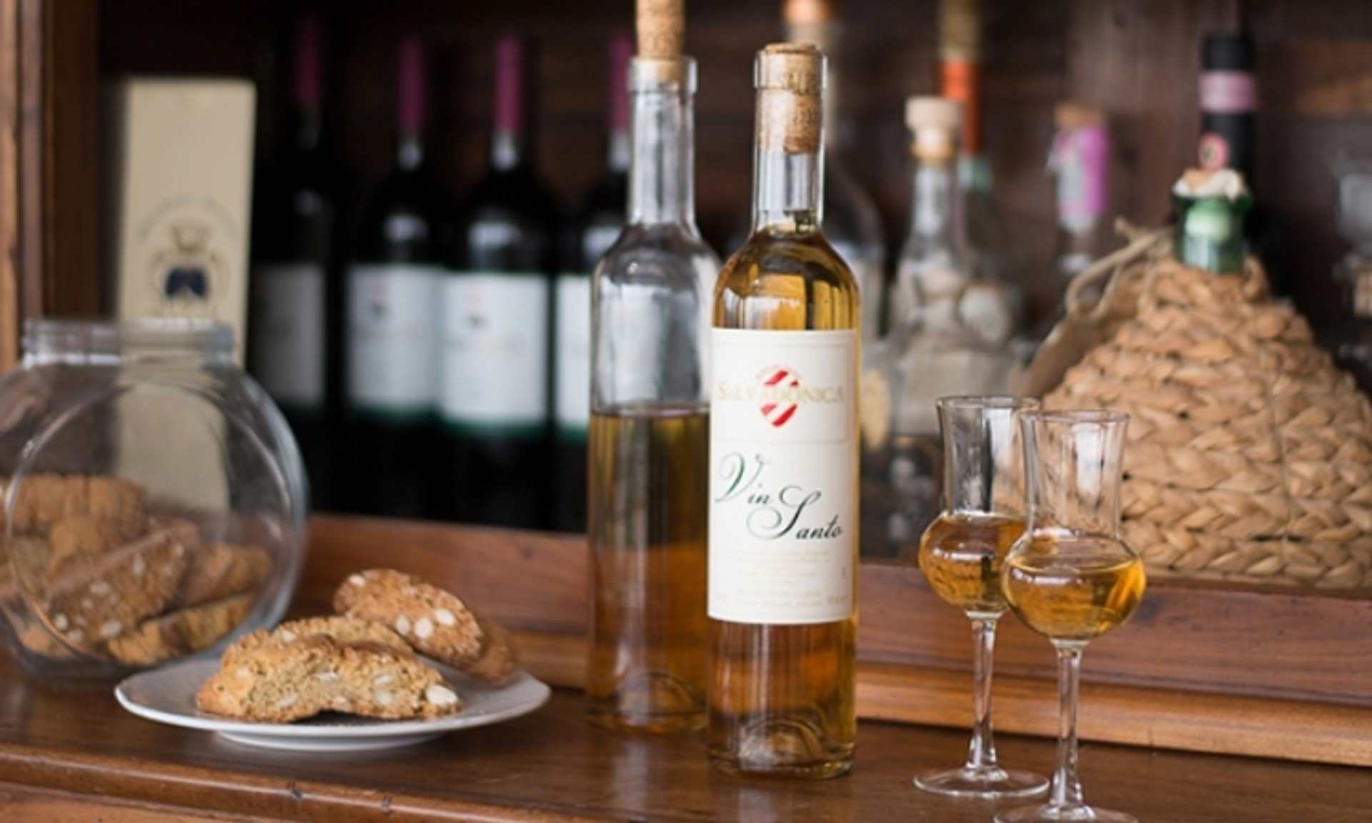The image depicts a wooden table with various wine bottles, glasses, and a plate of what appears to be biscuits or bread.