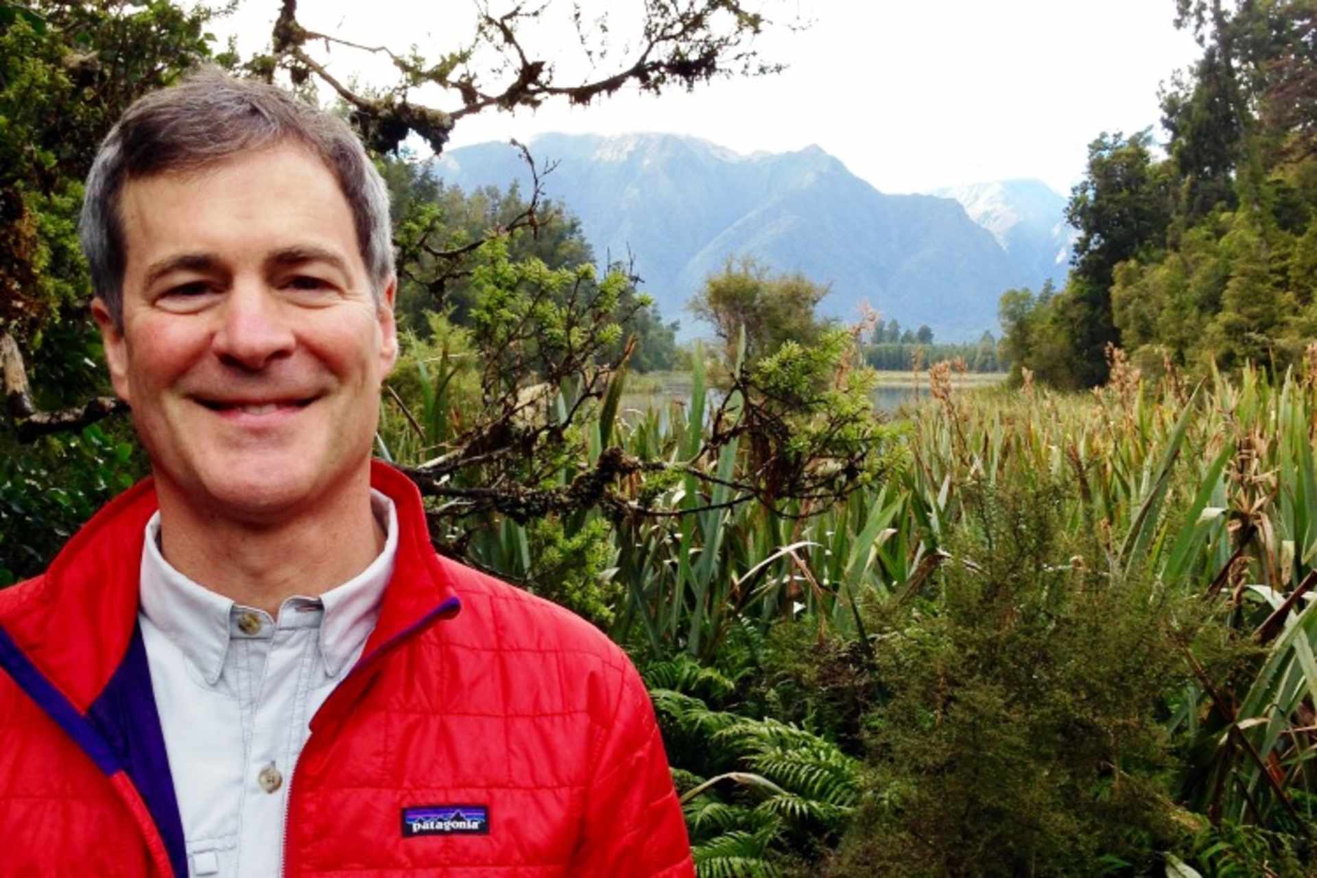 A smiling man wearing a red jacket stands in a lush, green landscape with mountains visible in the background.