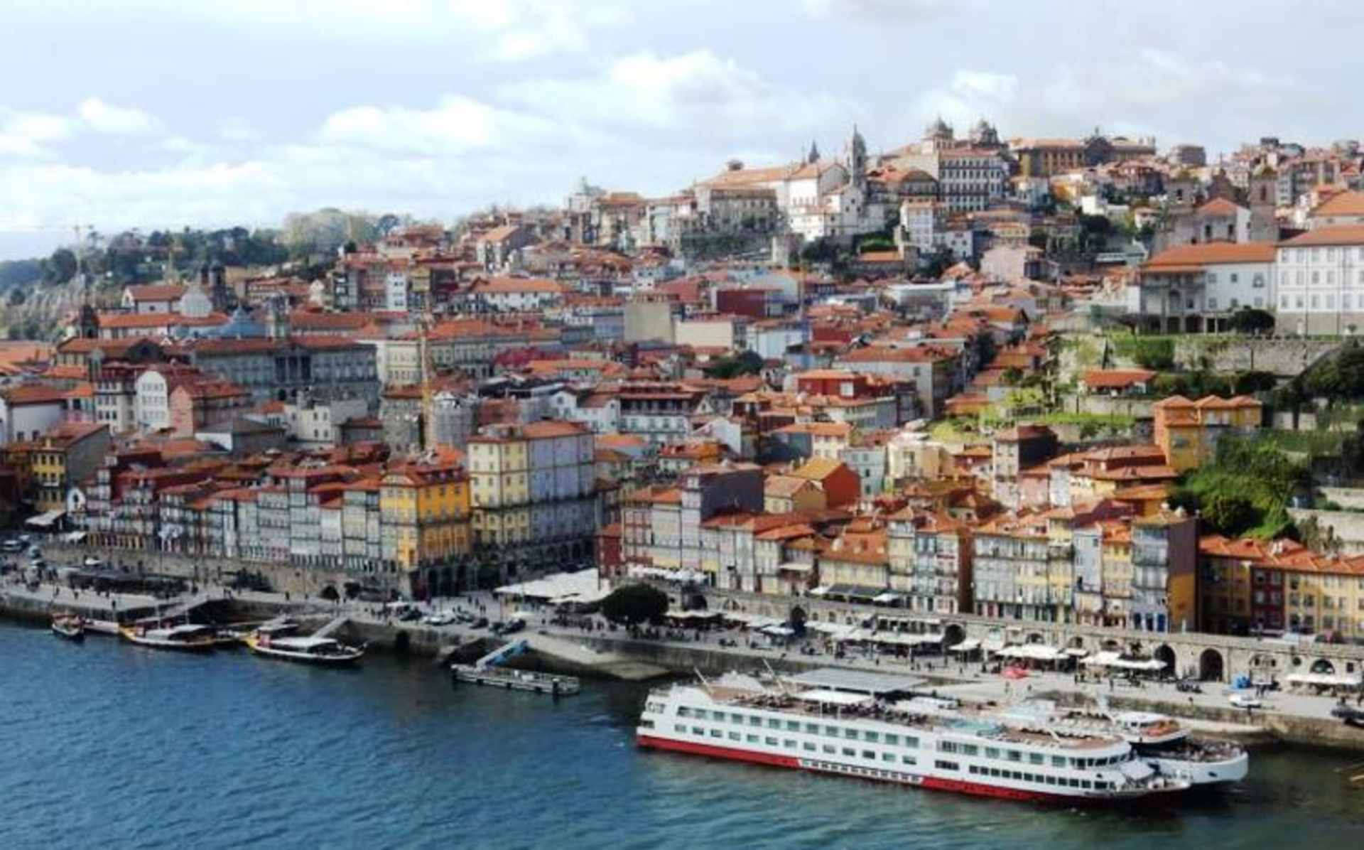 A vibrant cityscape with colorful buildings cascading down a hillside, overlooking a river with boats and ships docked along the waterfront.