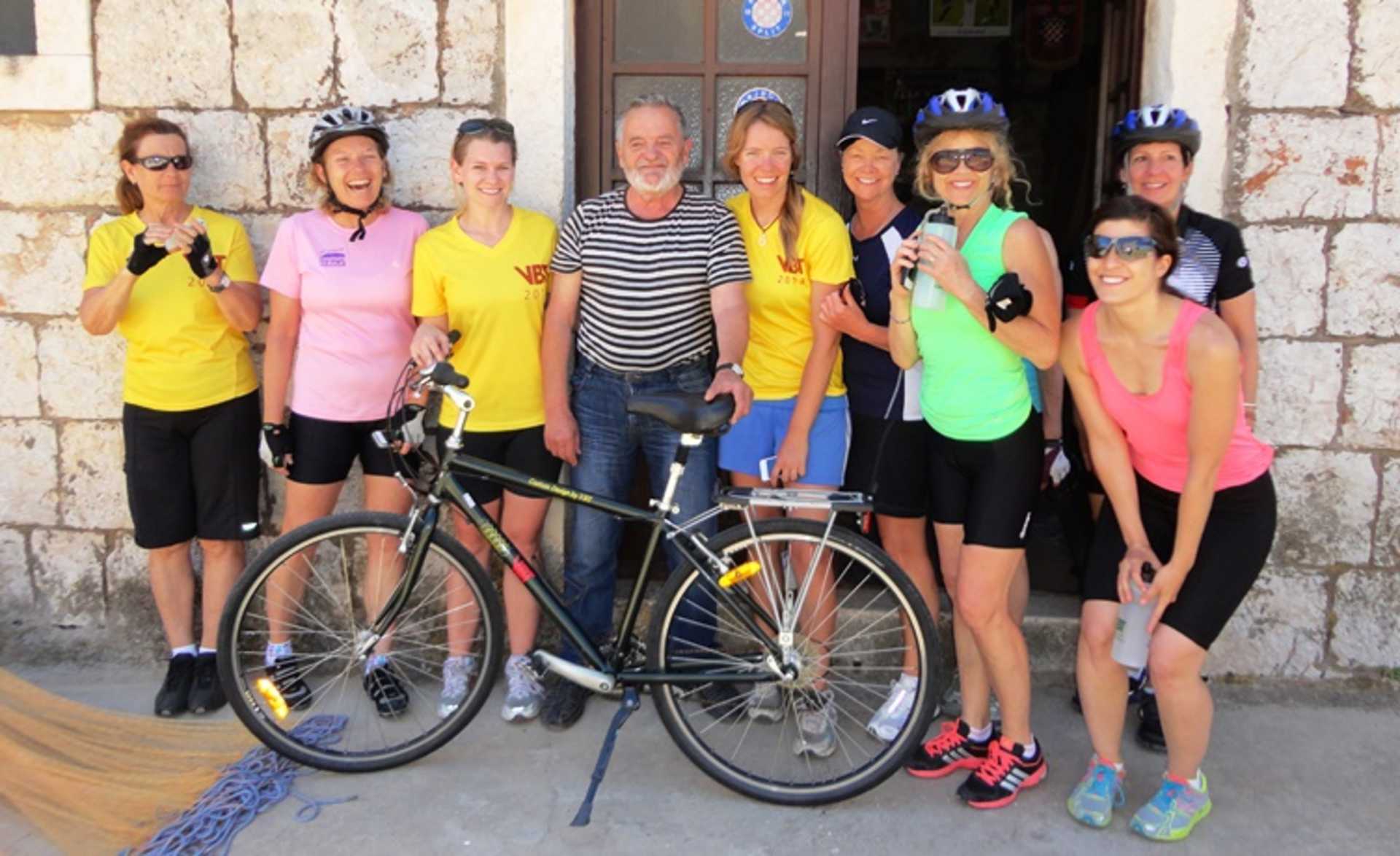 A group of people wearing colorful cycling gear, including jerseys, shorts, and helmets, posing together with a bicycle in front of a stone building.