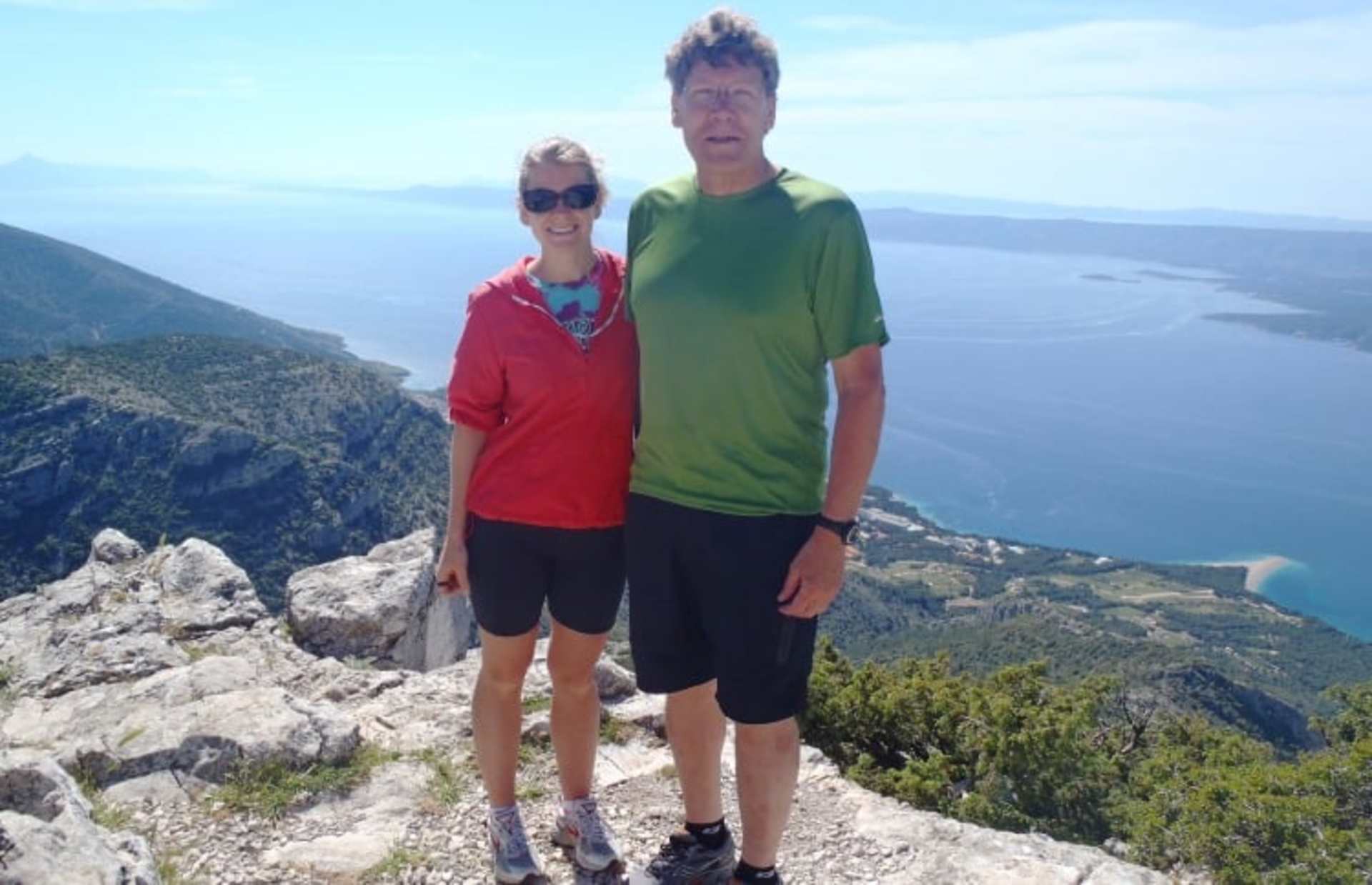Two people, a man and a woman, are standing on a rocky cliff overlooking a scenic coastal landscape with mountains and a body of water in the background.