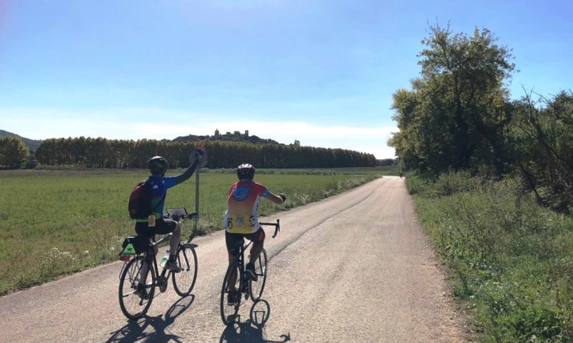Two cyclists riding on a dirt path through a grassy field, with a forested area and a building visible in the background.