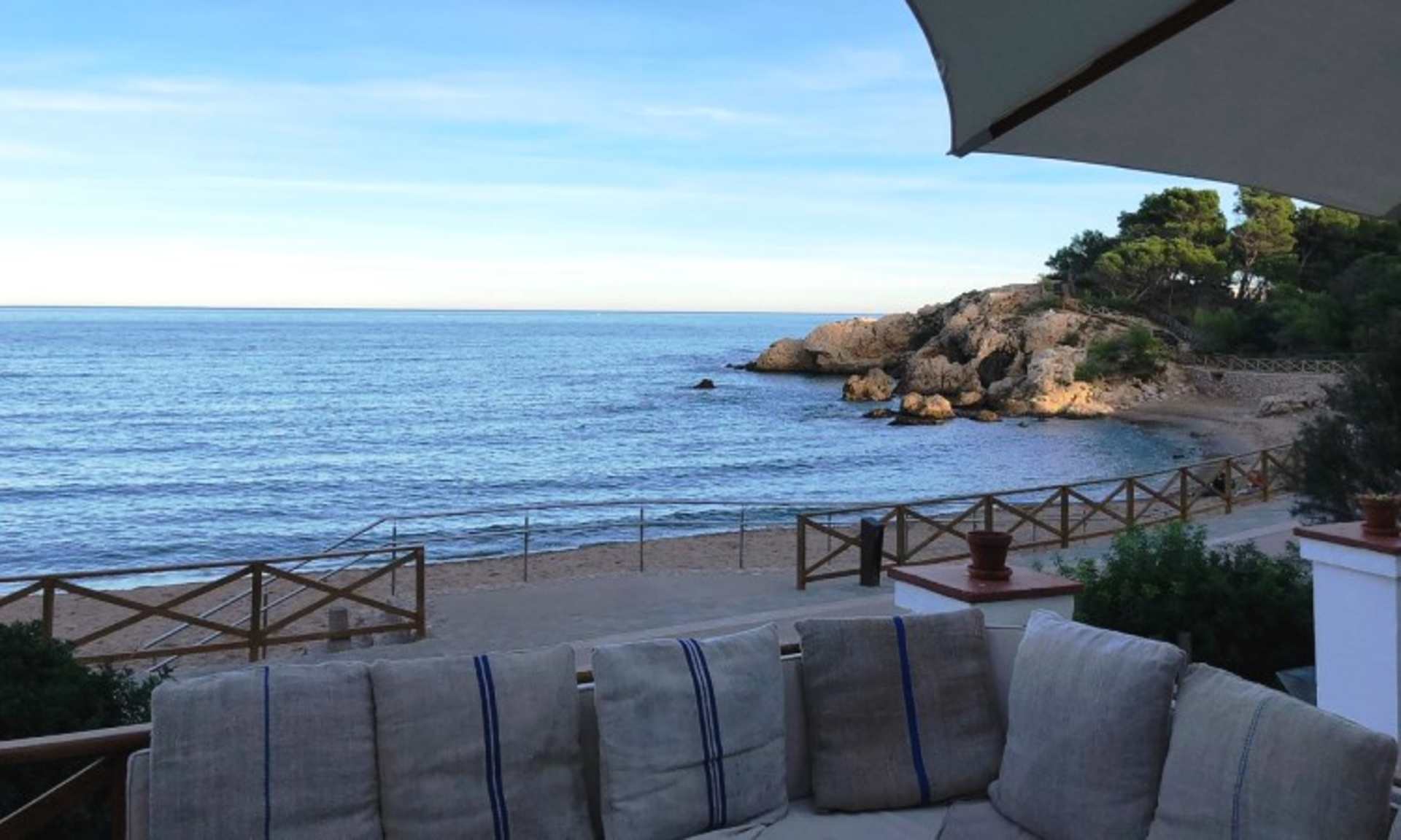 A scenic coastal view with a wooden railing and cushioned seating area in the foreground, overlooking a rocky shoreline and the calm, blue waters of the sea in the background.