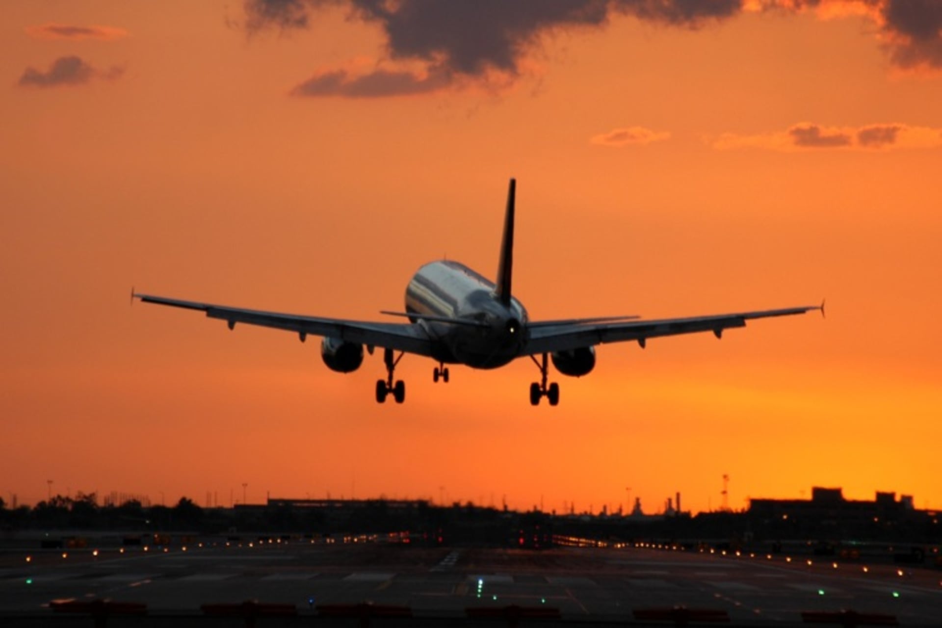 A commercial passenger aircraft is seen descending against a vibrant orange and cloudy sky, with the silhouette of a city skyline visible in the background.