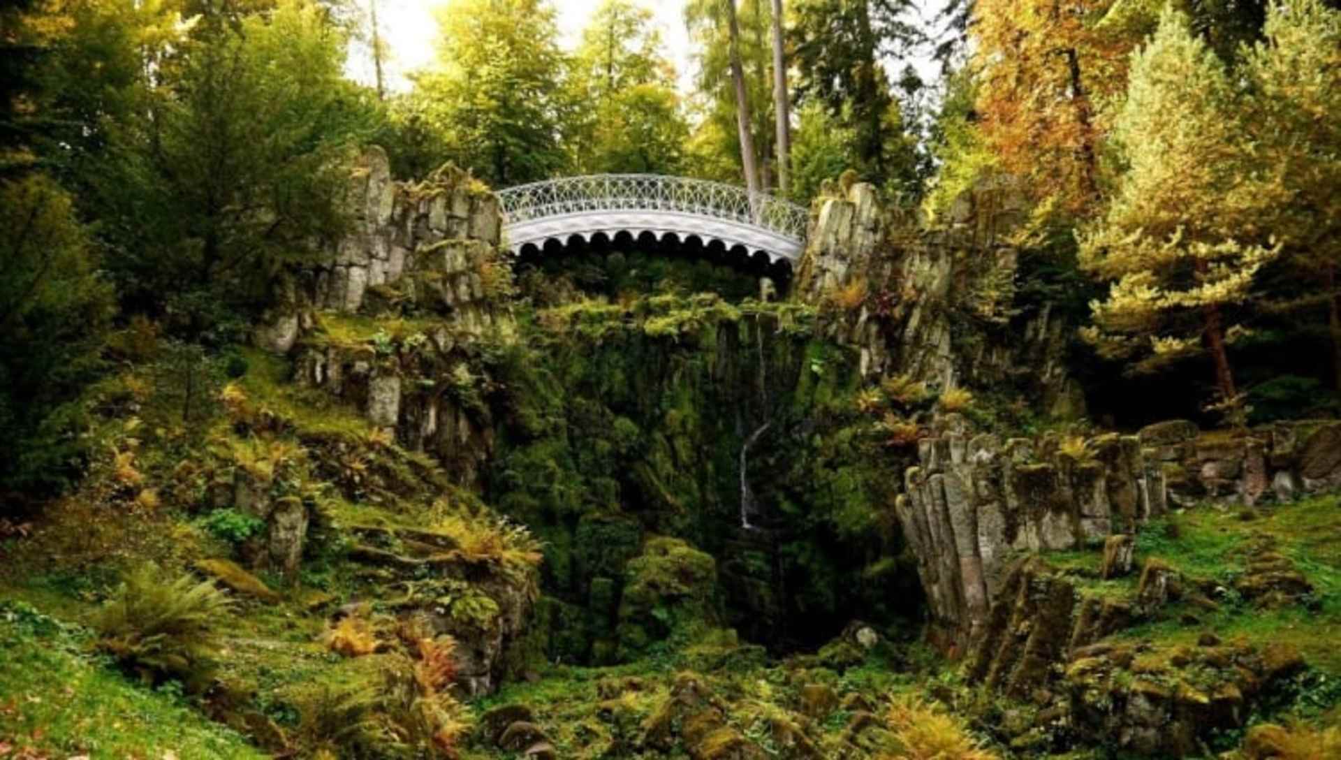 A lush, verdant forest landscape with a decorative stone bridge arching over a ravine, surrounded by vibrant foliage and towering trees.