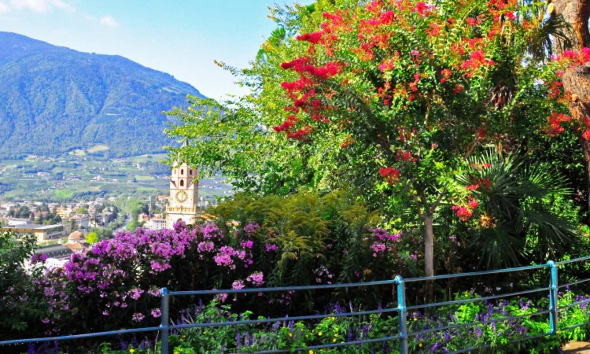 A lush, vibrant garden with blooming flowers and trees, overlooking a scenic town nestled among the mountains in the background.