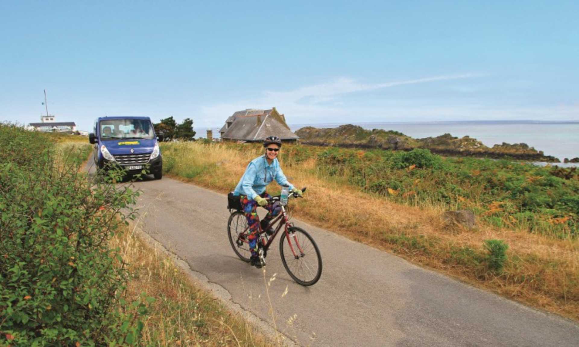 A person on a bicycle rides along a dirt path surrounded by lush vegetation, with a tractor and a thatched-roof building visible in the background.