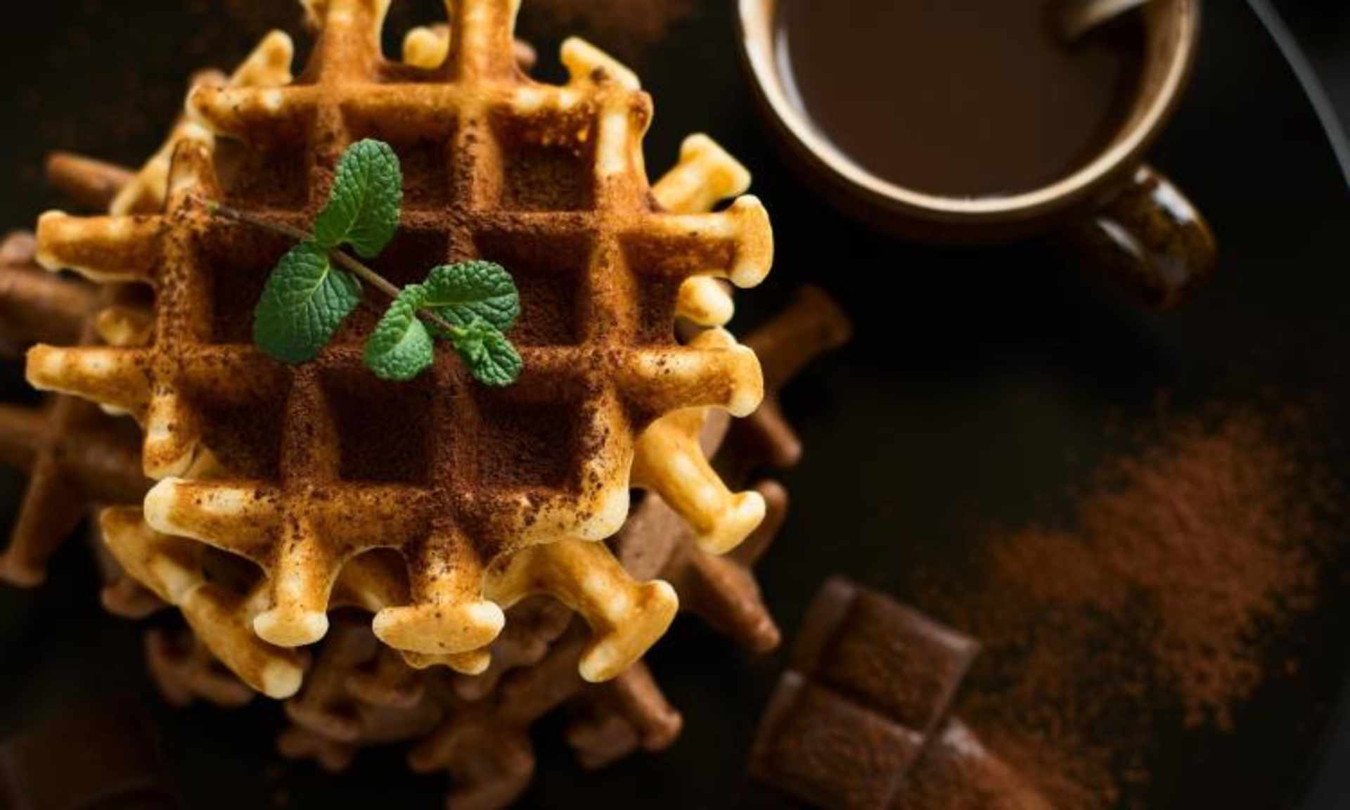 A golden-brown waffle topped with melted chocolate and fresh mint leaves, set against a dark background.