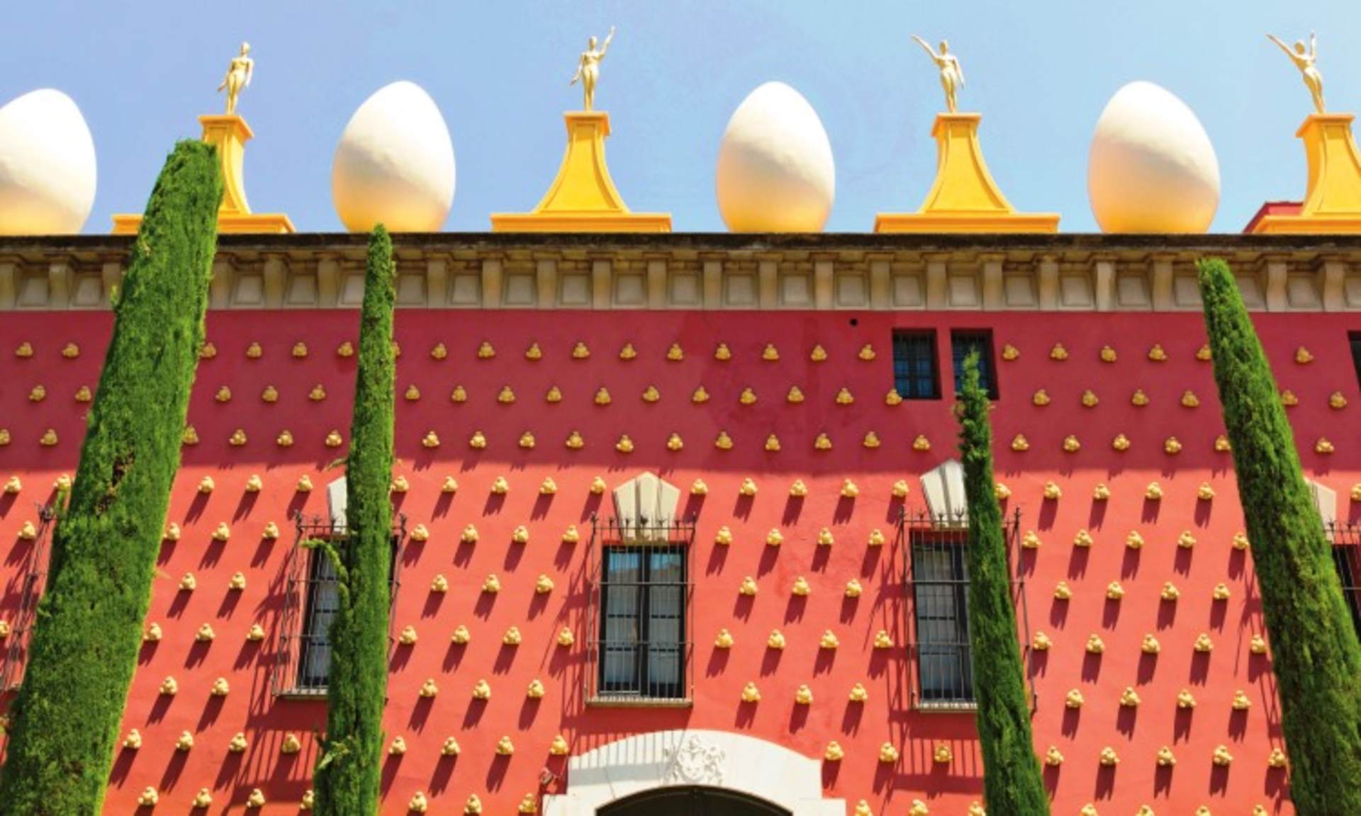A vibrant, red-colored building with golden spires and cypress trees in the foreground, set against a clear blue sky.
