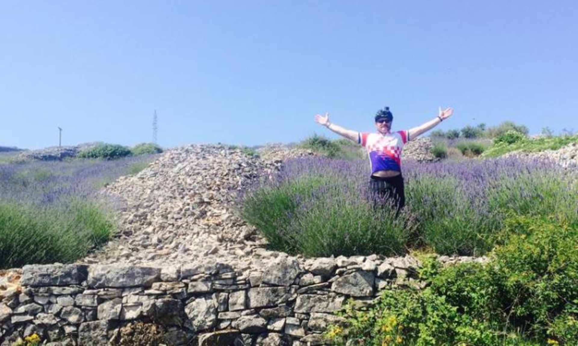 A person stands with arms raised in a field of lavender, surrounded by a stone wall and a rural landscape in the background.