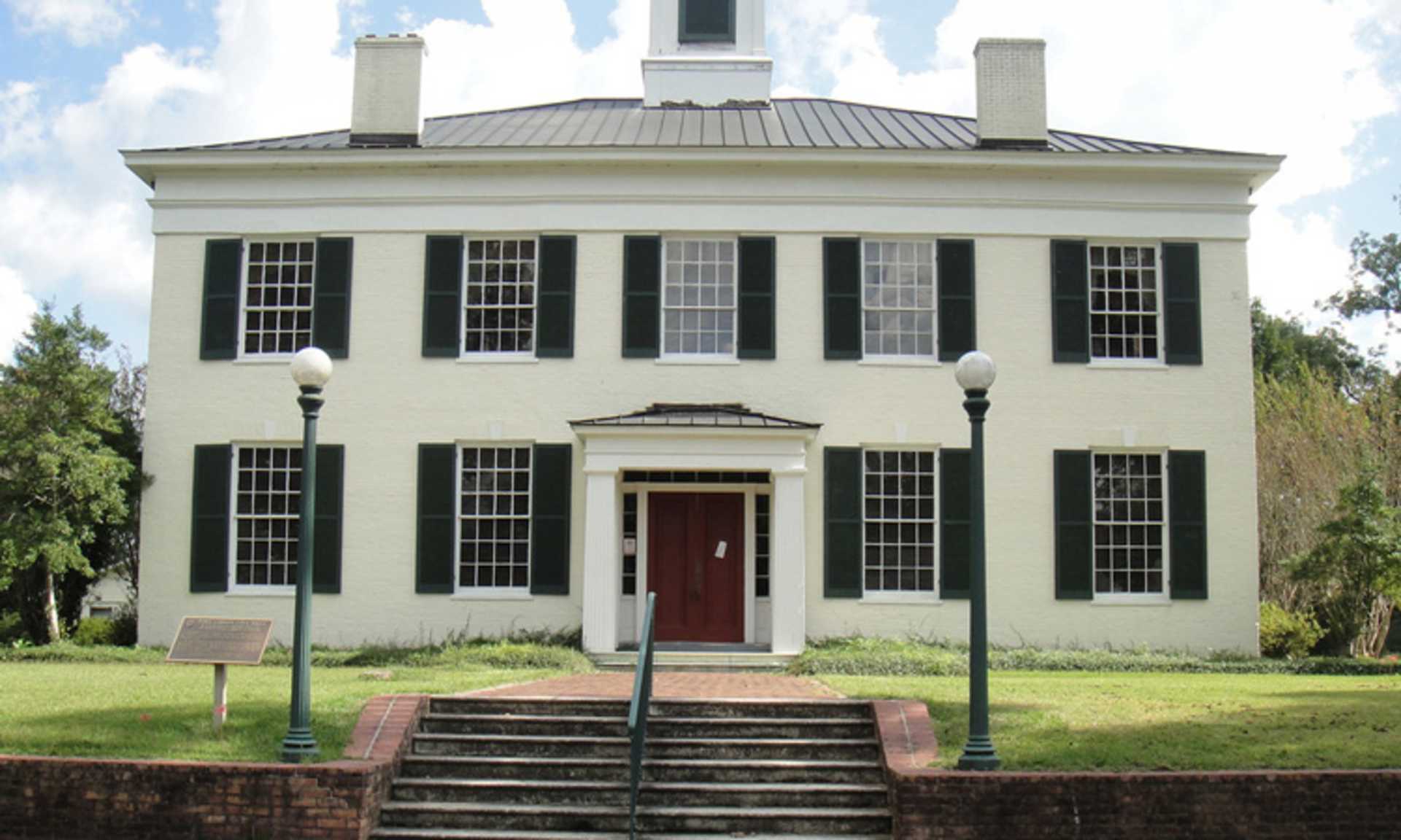 A stately white colonial-style building with a red door and black shutters stands on a grassy lawn, surrounded by trees and a clear blue sky.