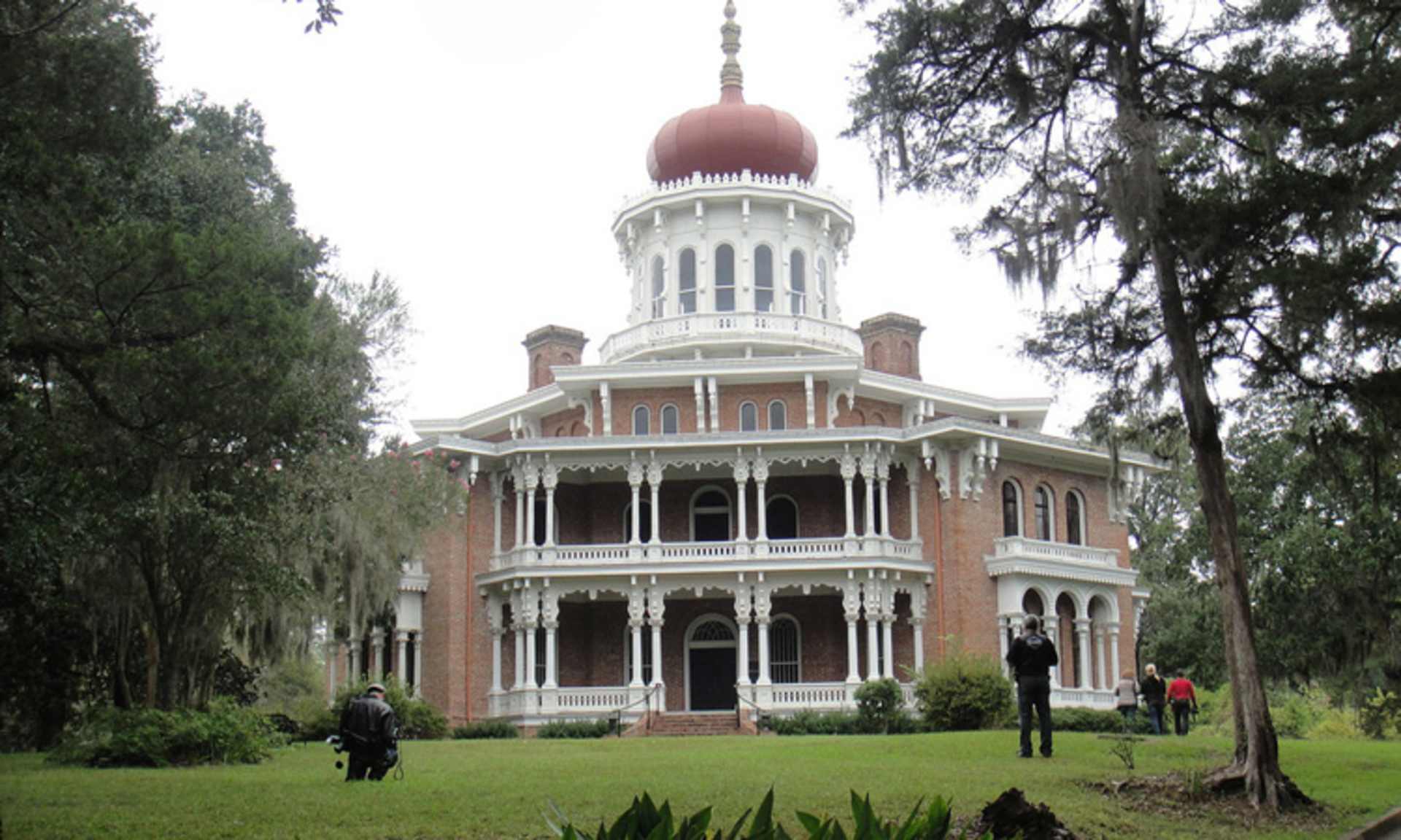 A grand, ornate Victorian-style mansion with a prominent domed roof stands amidst a lush, verdant landscape, surrounded by towering trees and a grassy lawn where a few people can be seen walking.