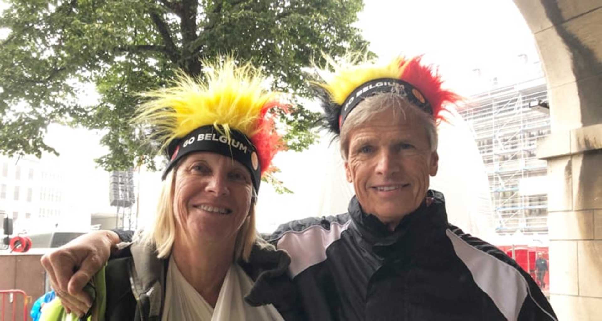 Two people wearing colorful headdresses with feathers, smiling and posing in front of a background of trees and buildings.