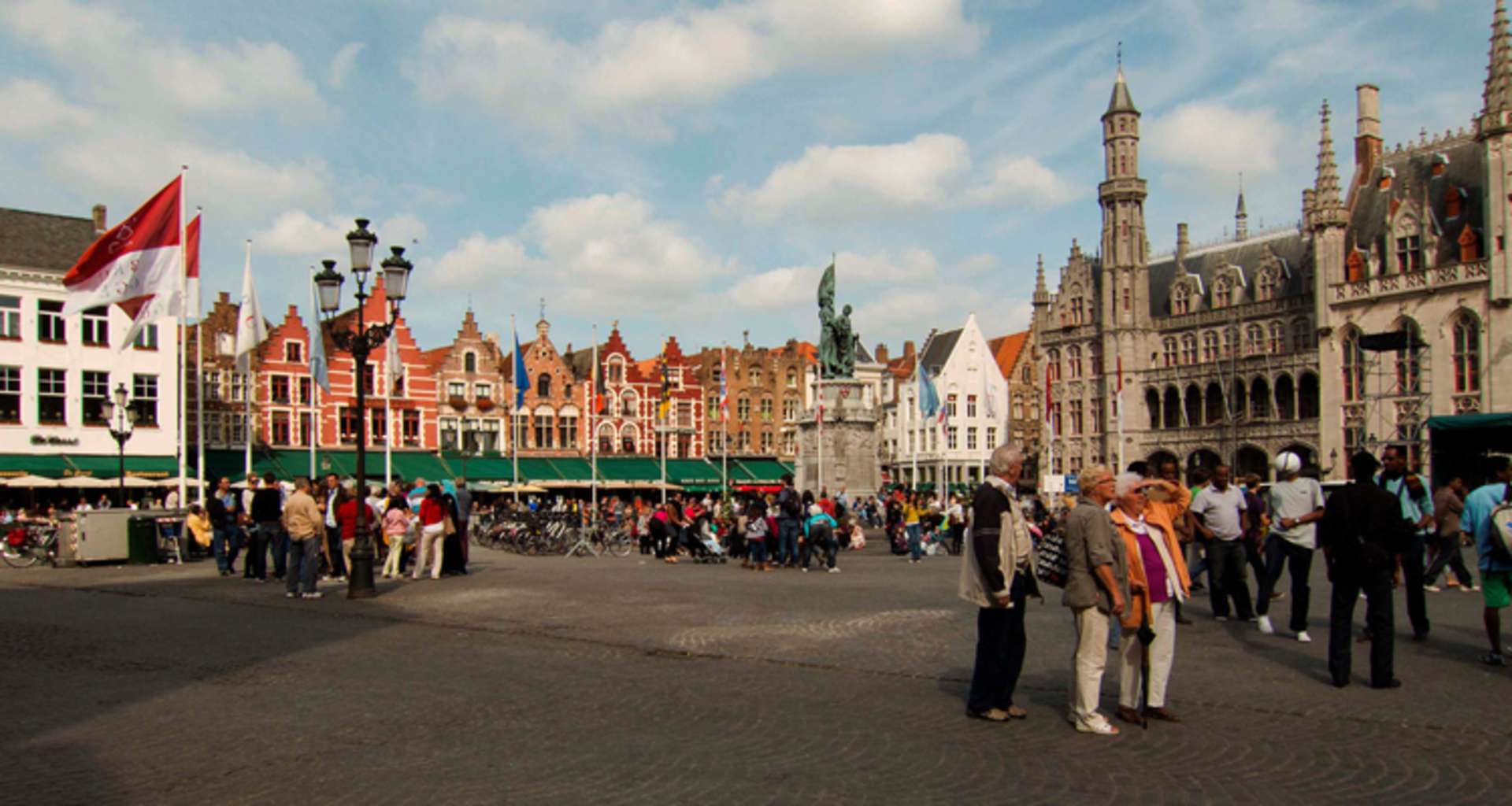 A bustling town square with colorful historic buildings, flags, and a crowd of people walking through the cobblestone streets.