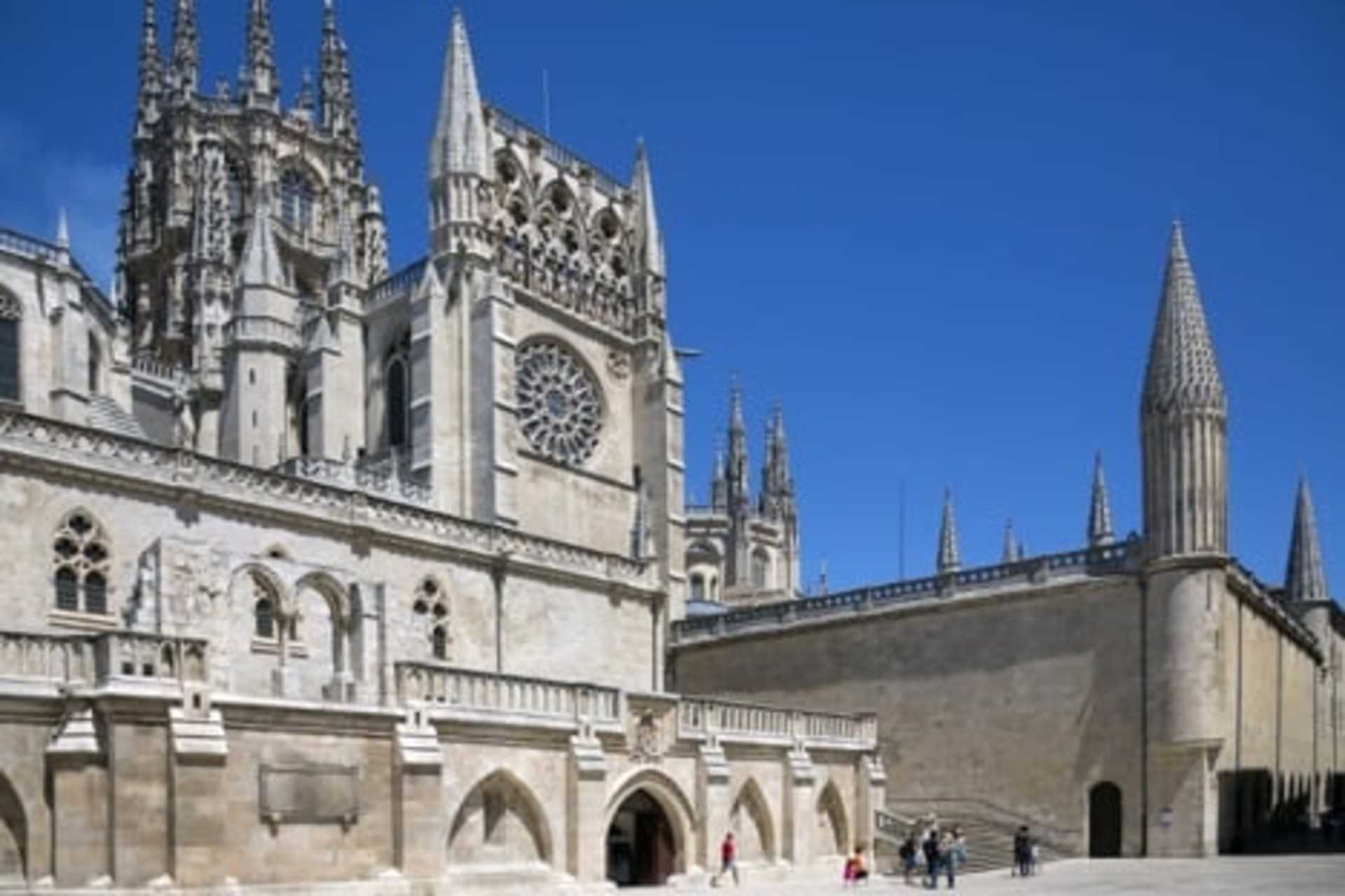 A grand, ornate cathedral with intricate Gothic architecture stands tall against a clear blue sky, with people walking along the steps in the foreground.
