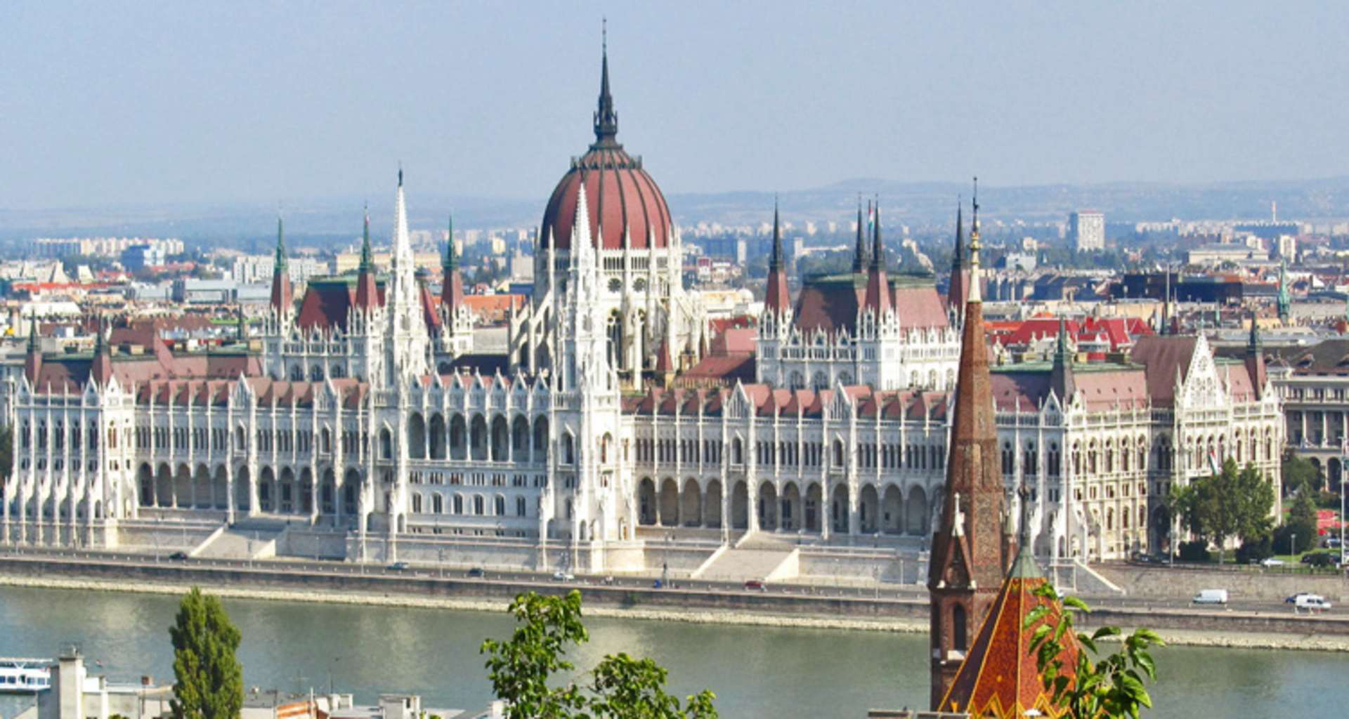 The image depicts the iconic Hungarian Parliament Building situated along the Danube River, with the city skyline of Budapest visible in the background.
