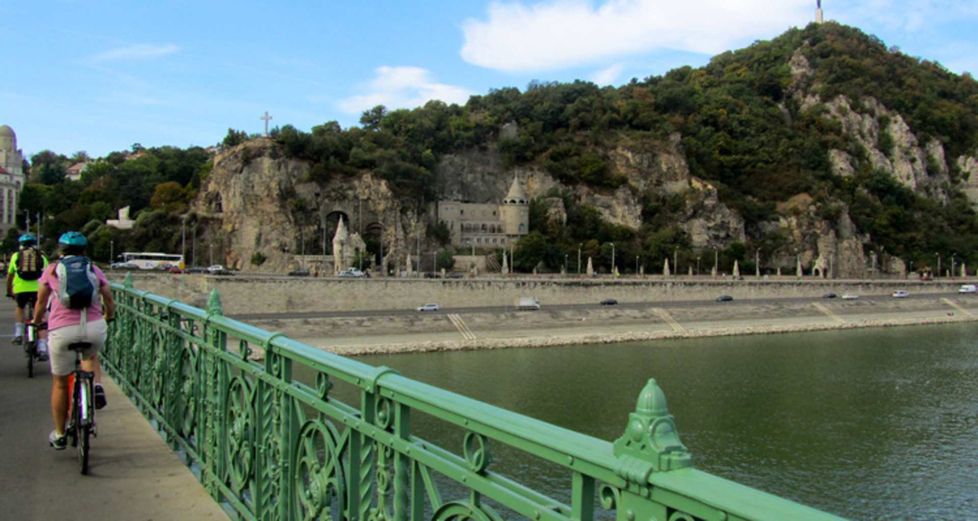 A scenic view of a river surrounded by towering cliffs and lush greenery, with a green pedestrian bridge in the foreground.