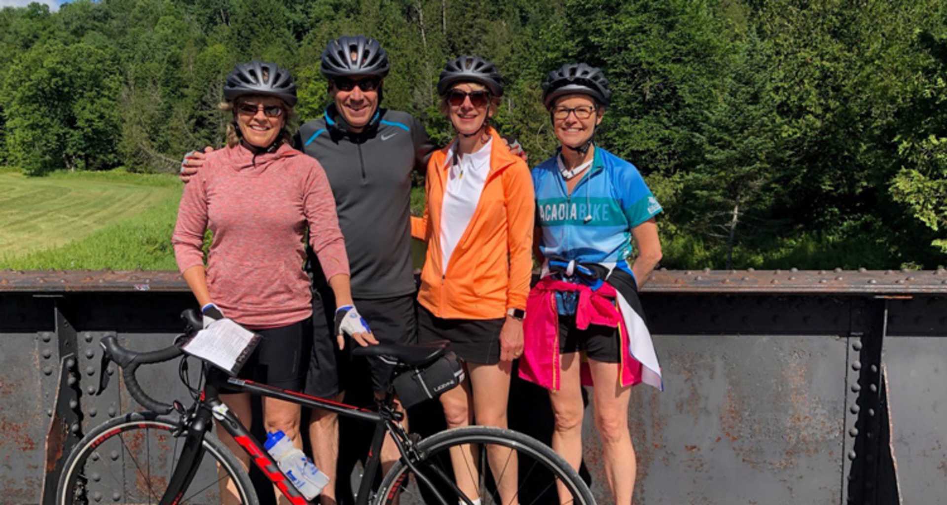 A group of four cyclists, wearing colorful cycling gear, posing together in front of a wooded area with a grassy field in the background.