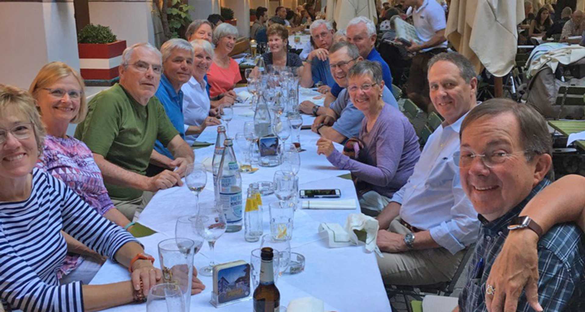 A group of people, mostly elderly, seated around a long table enjoying a meal together in what appears to be an outdoor setting with buildings and greenery visible in the background.