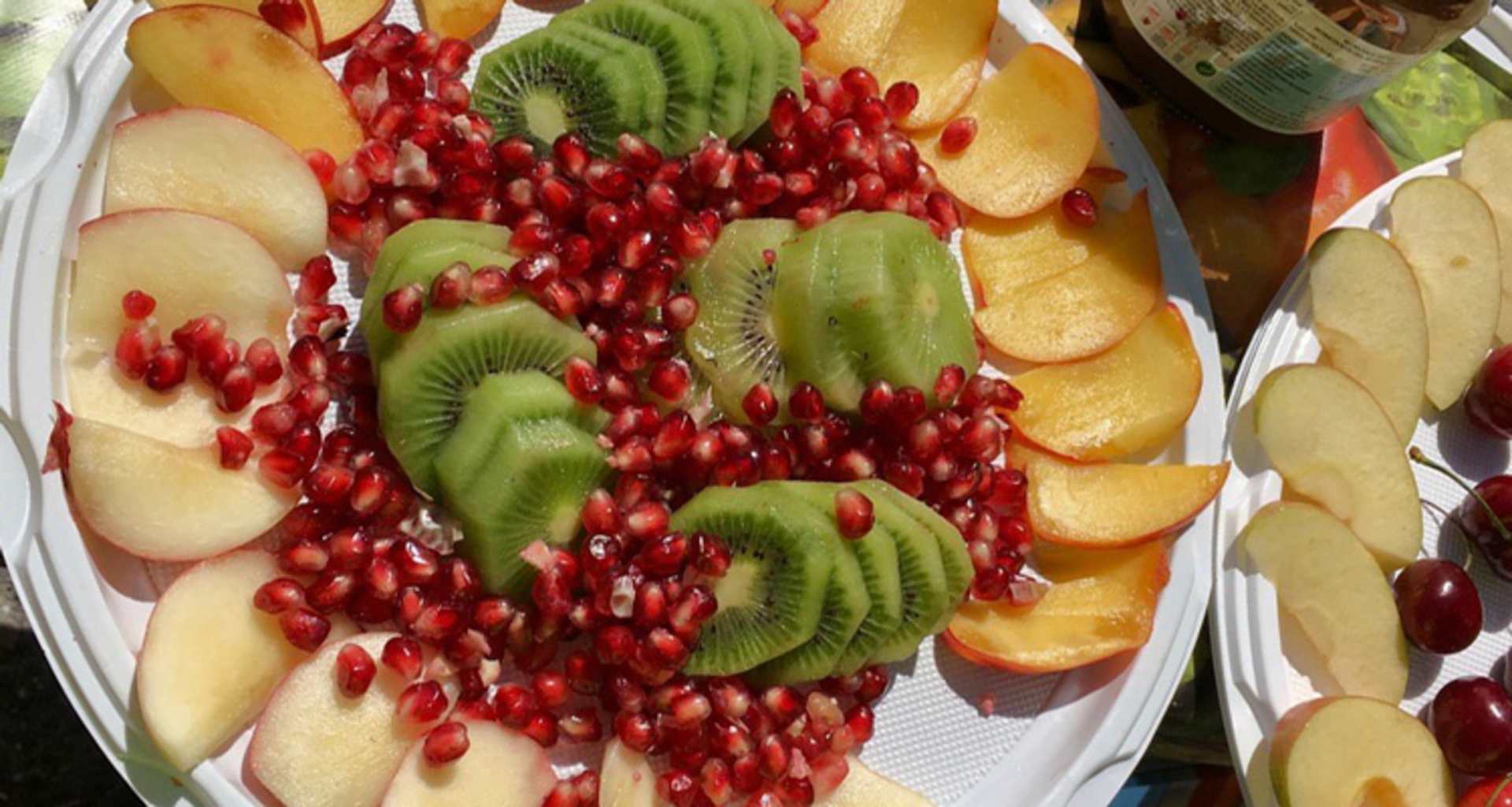 A colorful and vibrant assortment of various fruits, including sliced kiwi, pomegranate seeds, and peach or nectarine slices, arranged on a white platter or plate.