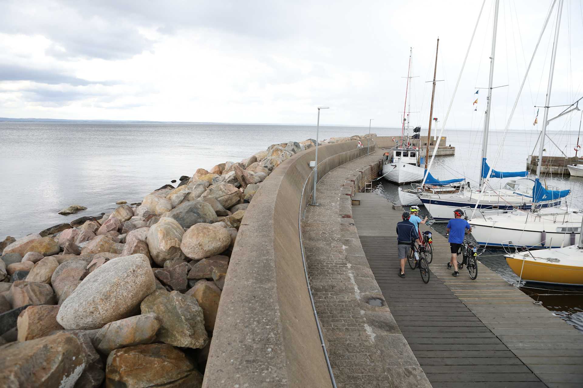 A wooden walkway leads along a rocky shoreline, with sailboats and yachts moored in the calm waters of the harbor in the background.