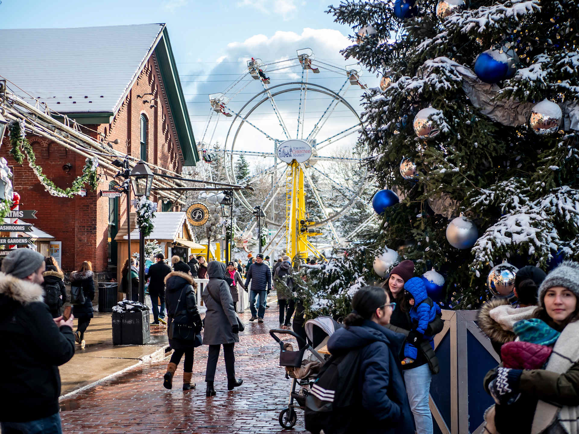 A snowy winter scene with a bustling crowd walking along a street lined with festive decorations, including a large Ferris wheel in the background.