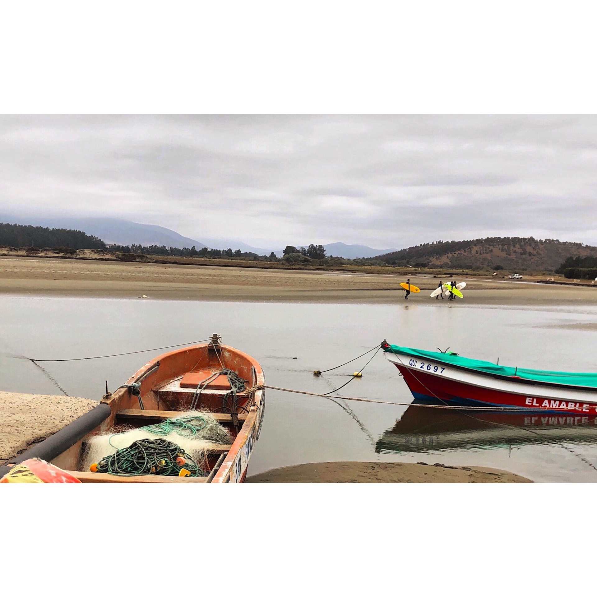 The image depicts a serene lakeside scene with several small boats moored in the foreground, surrounded by a hilly, forested landscape in the background.