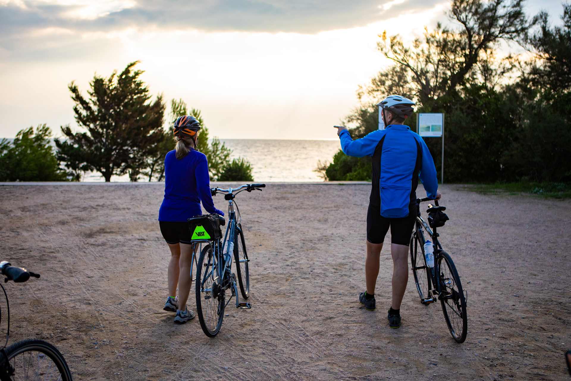 Two cyclists, one in a blue jersey and the other in a blue jacket, stand with their bicycles on a dirt path surrounded by trees and a body of water in the background.