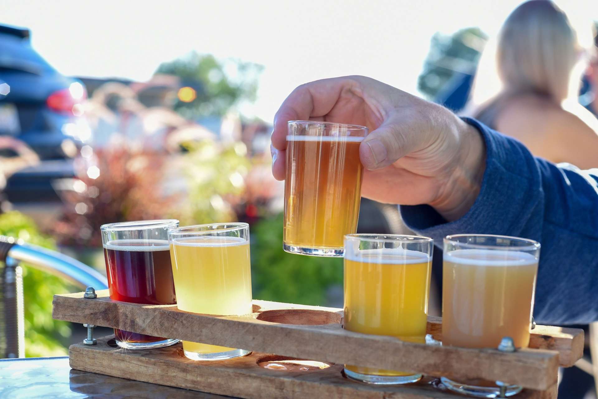 A hand holding a glass of amber-colored liquid, surrounded by other glasses of various colored beverages, set against a blurred background of people and outdoor scenery.