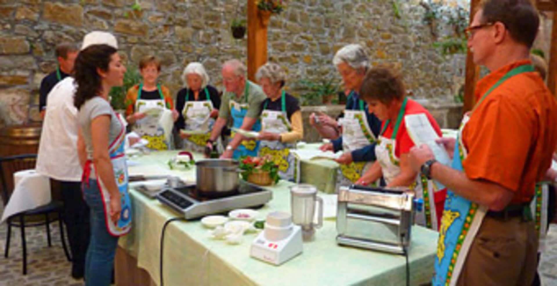 A group of people, dressed in colorful aprons, are gathered around a table in a rustic setting, engaged in what appears to be a cooking or food preparation activity.