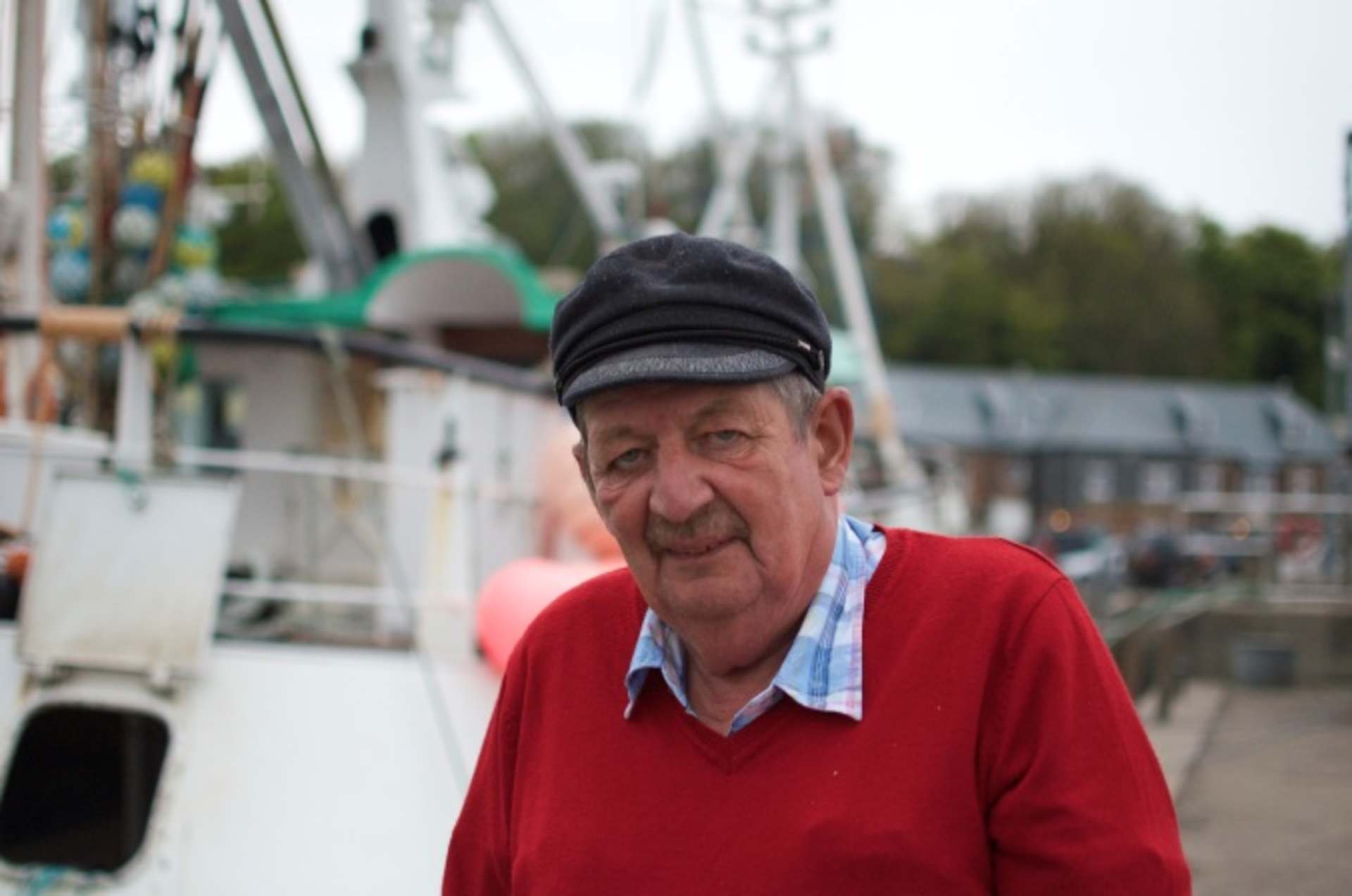 An elderly man wearing a red sweater and a black cap stands in front of a fishing boat in a harbor setting.