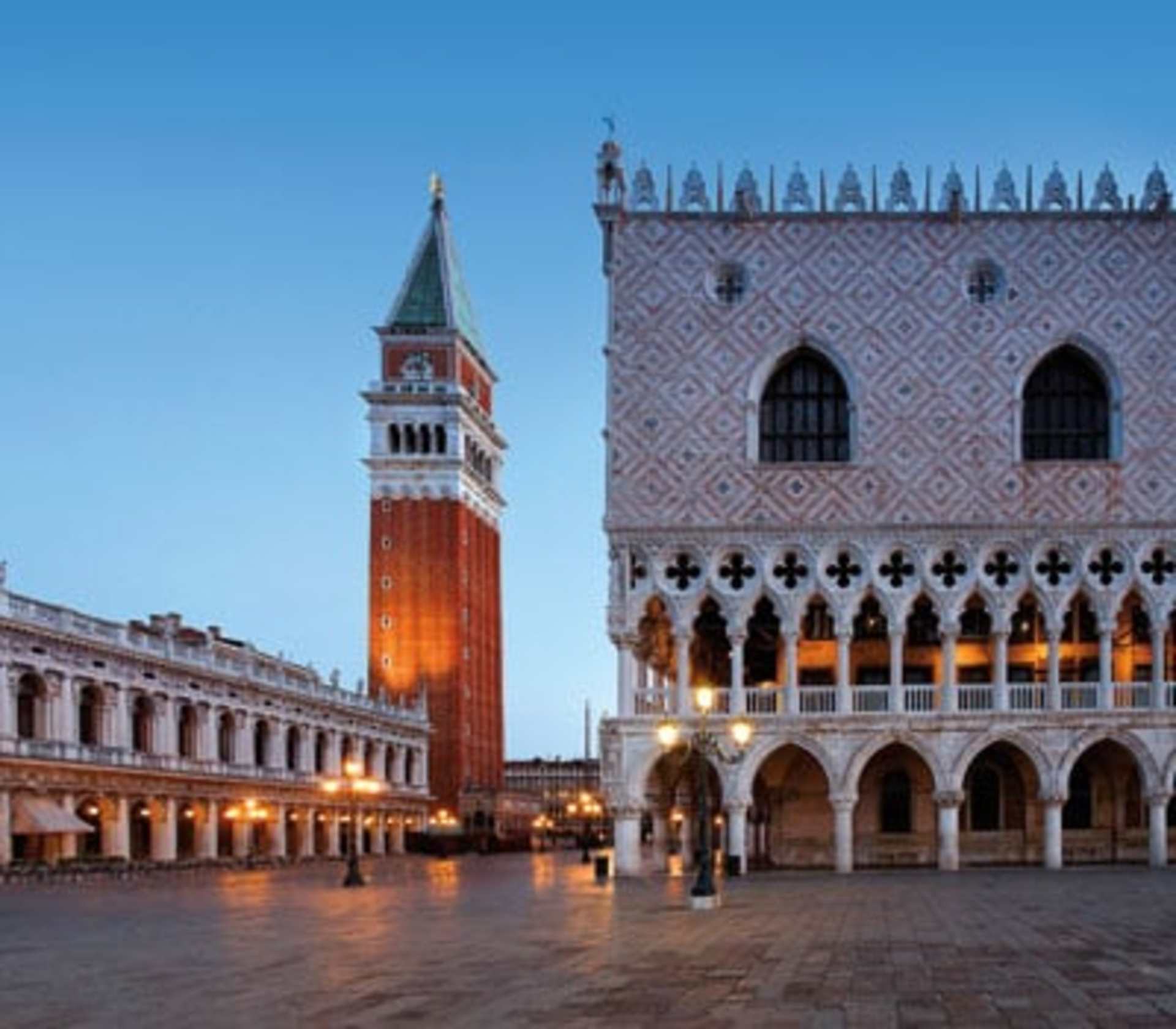 A grand, ornate palace with a tall bell tower stands in a large, open square, illuminated by the warm glow of lights as the sun sets in the background.
