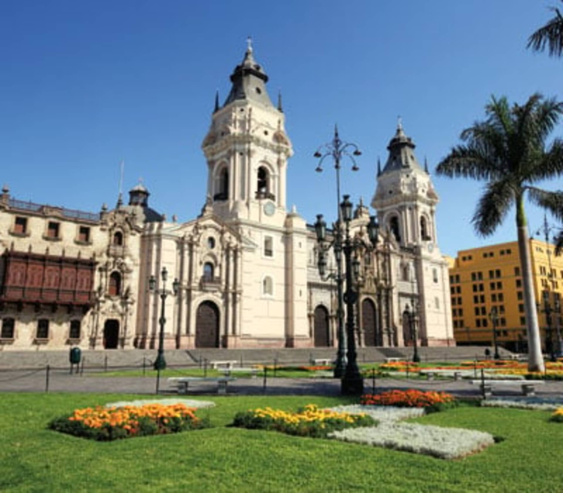 A grand, ornate cathedral with multiple domes and towers stands in the center of a well-manicured plaza, surrounded by lush greenery and colorful flower beds in the foreground.