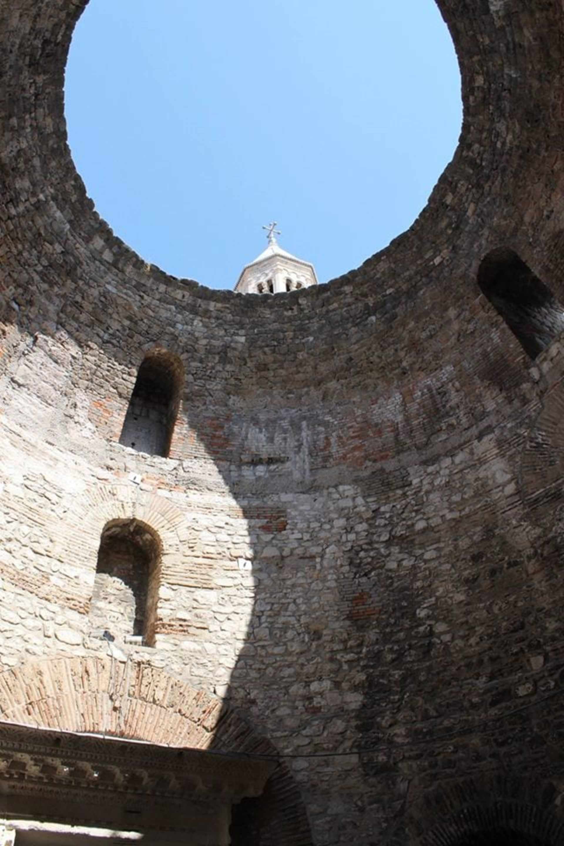 A circular stone structure with an opening in the center, revealing a small domed building in the background against a clear blue sky.