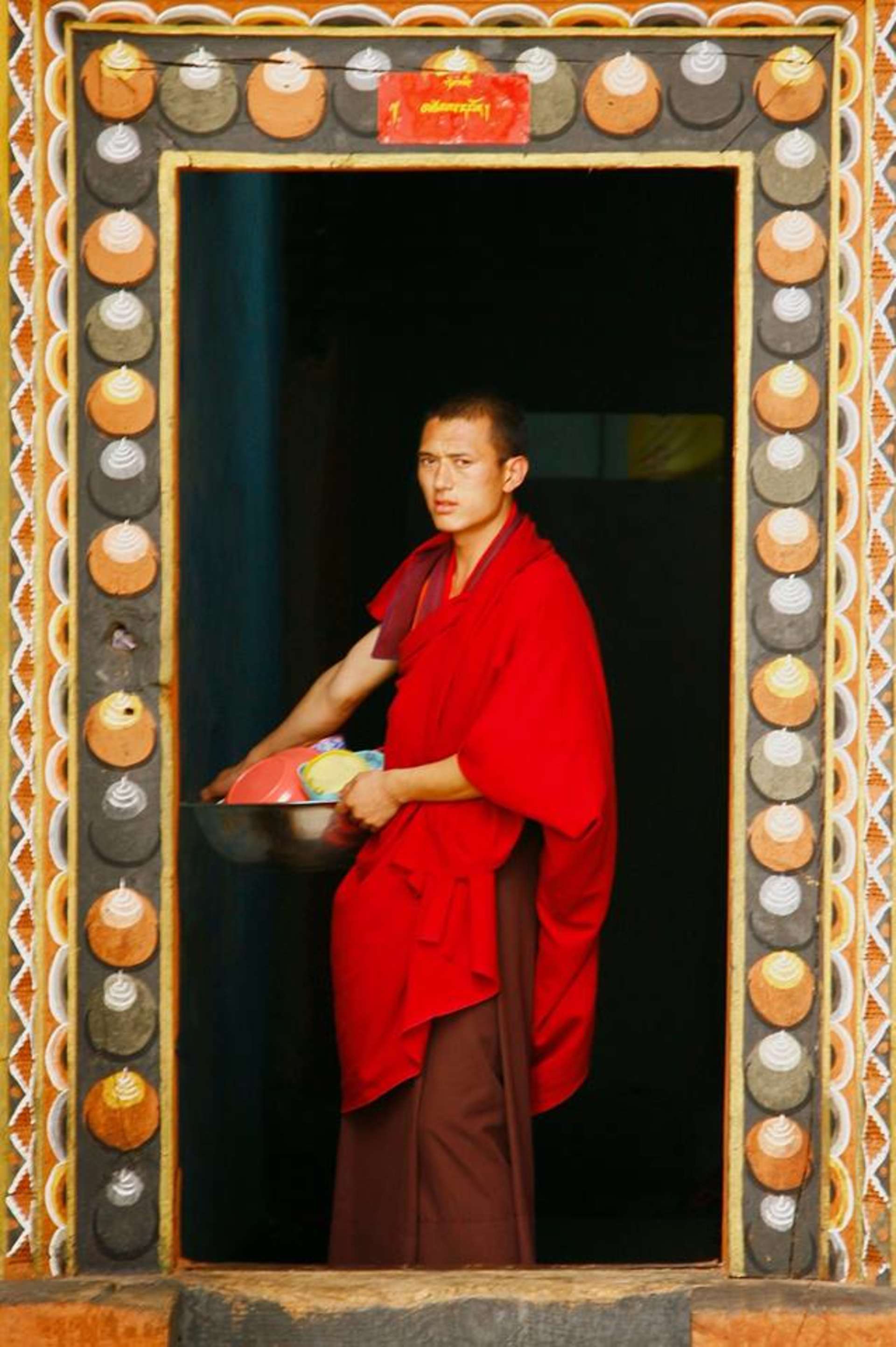 A person wearing a bright red robe stands in a doorway framed by an ornate, patterned border.