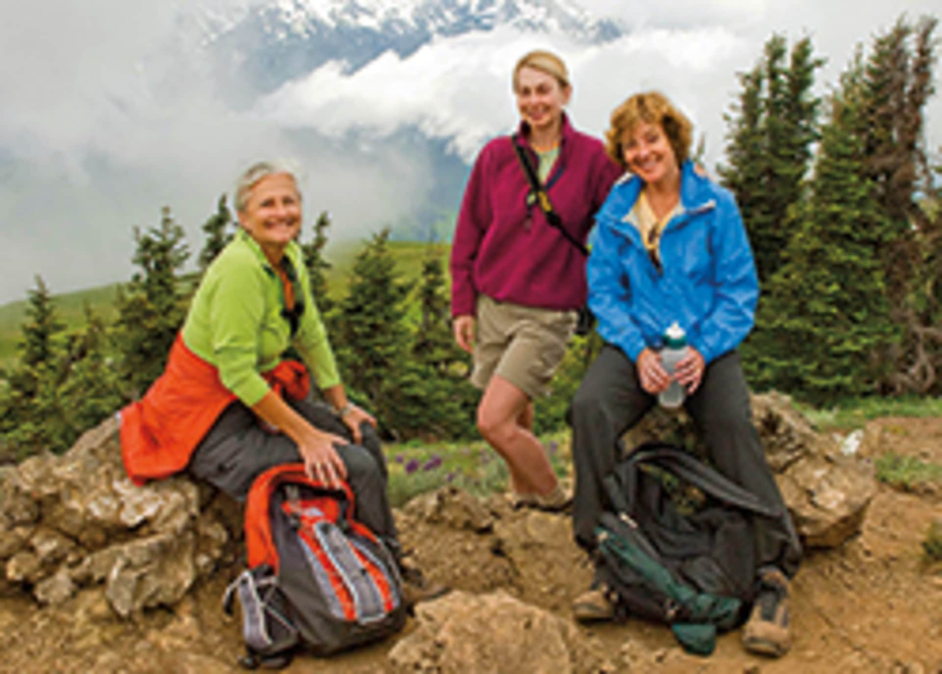 Three women in colorful outdoor clothing stand on a rocky hillside with a forested mountain range in the background.