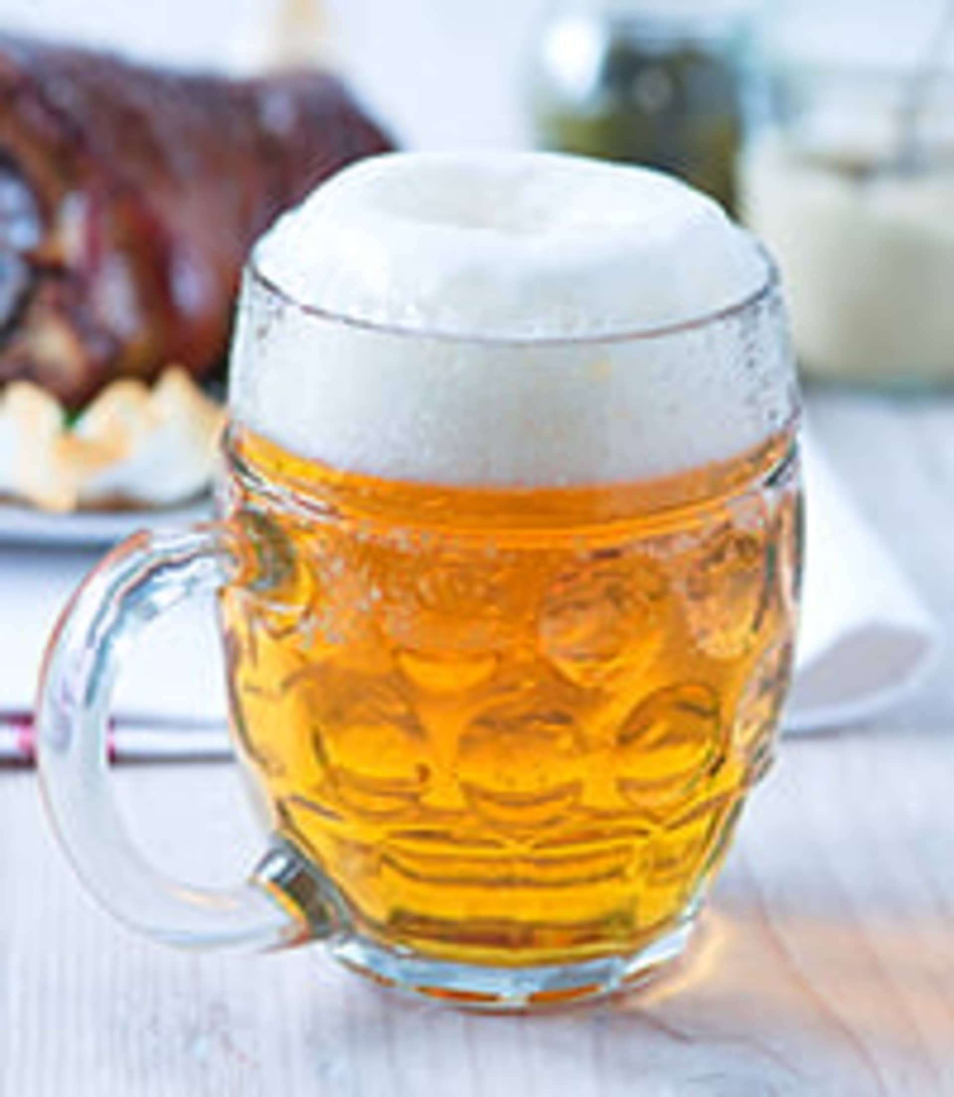 A glass mug filled with golden-colored beer sits on a wooden table, with a roasted meat dish visible in the background.