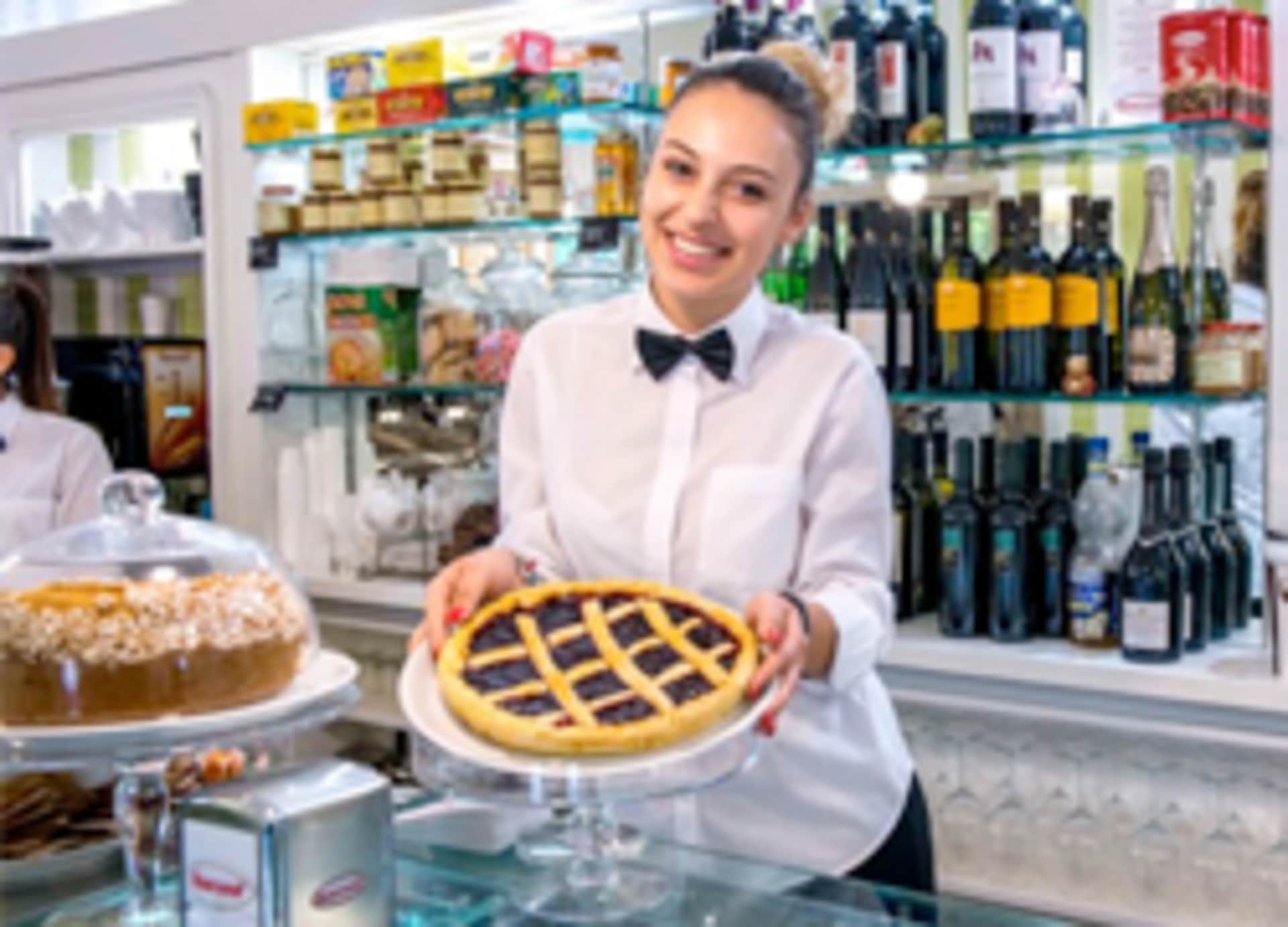 A smiling woman in a white shirt and bow tie is holding a freshly baked pie in a bakery or cafe setting, with shelves of wine bottles and other food items visible in the background.