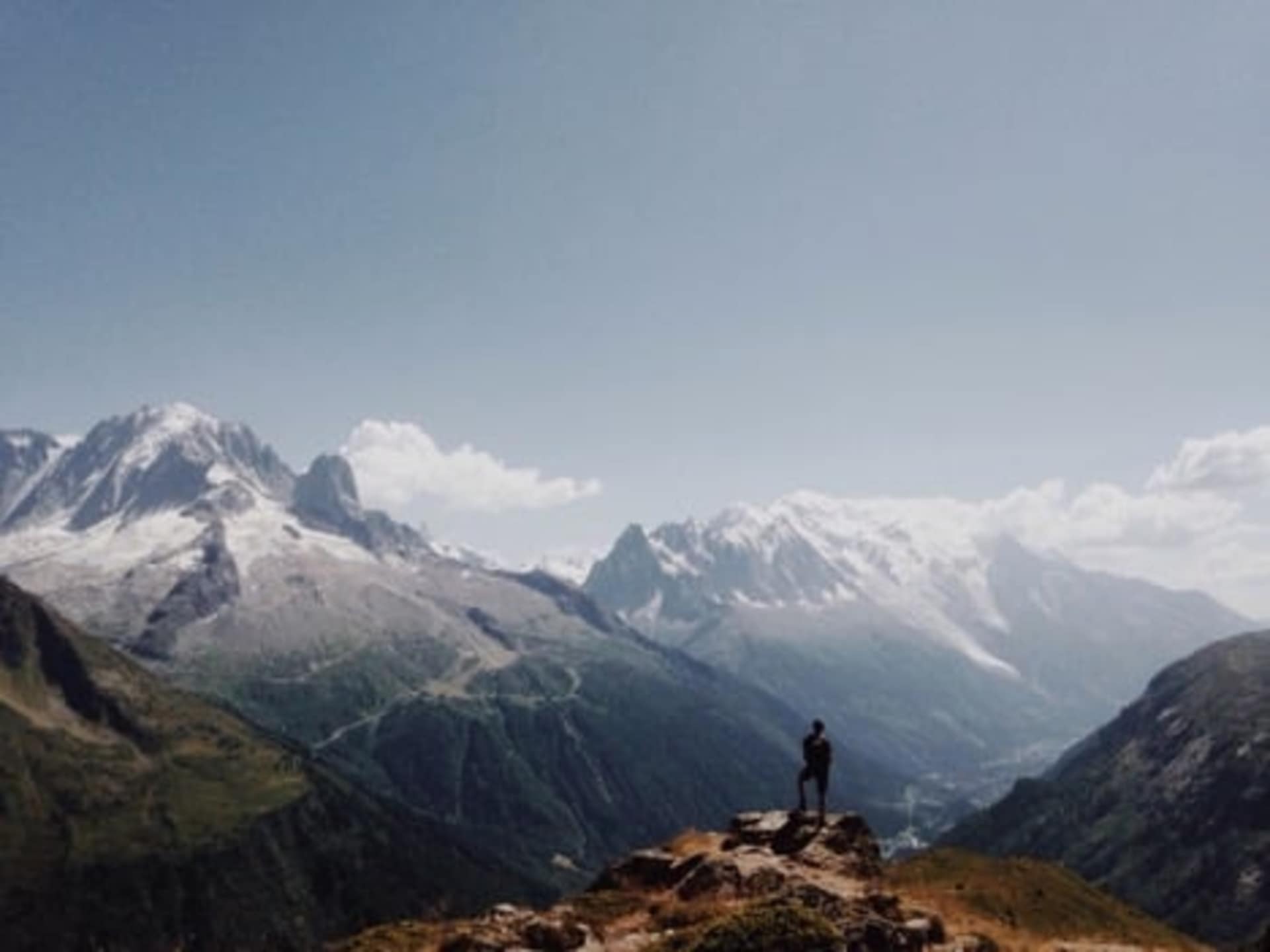 A lone person stands atop a rocky outcrop, gazing out at the majestic snow-capped mountains in the distance, surrounded by a vast, serene landscape.