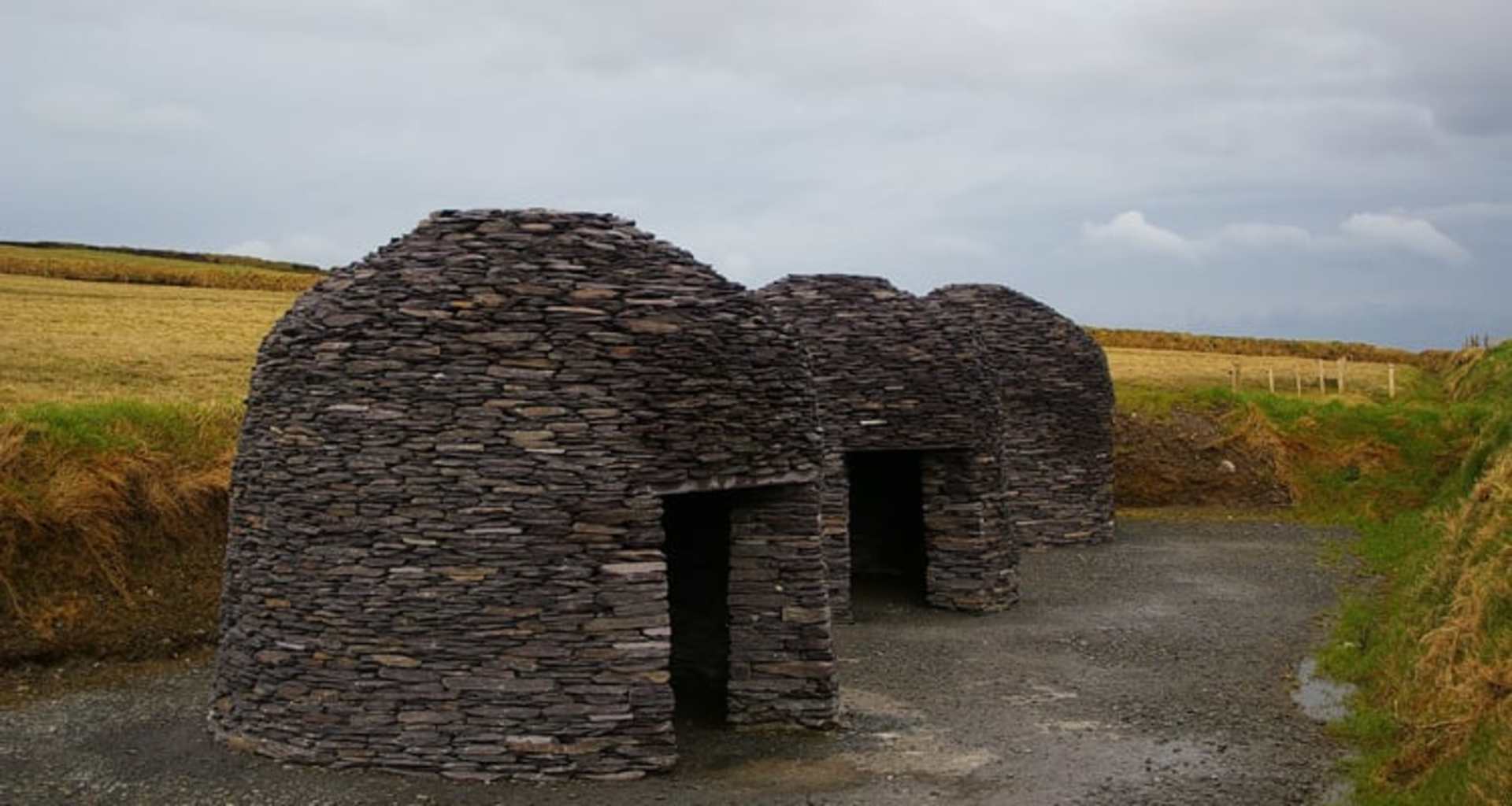 A stone structure with multiple domed roofs stands in a grassy field, with a dirt path leading towards it and a cloudy sky in the background.