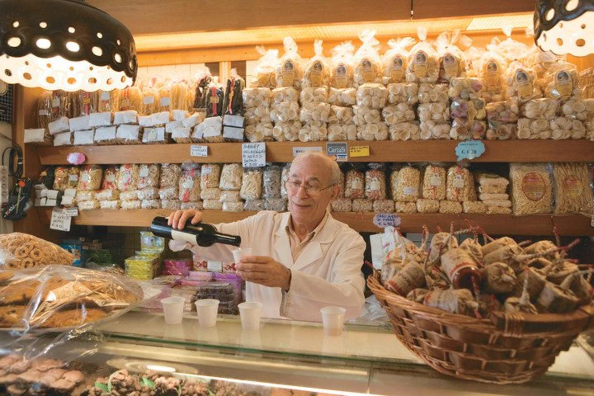 A well-stocked bakery counter with various baked goods, including breads and pastries, with a smiling elderly woman behind the counter.